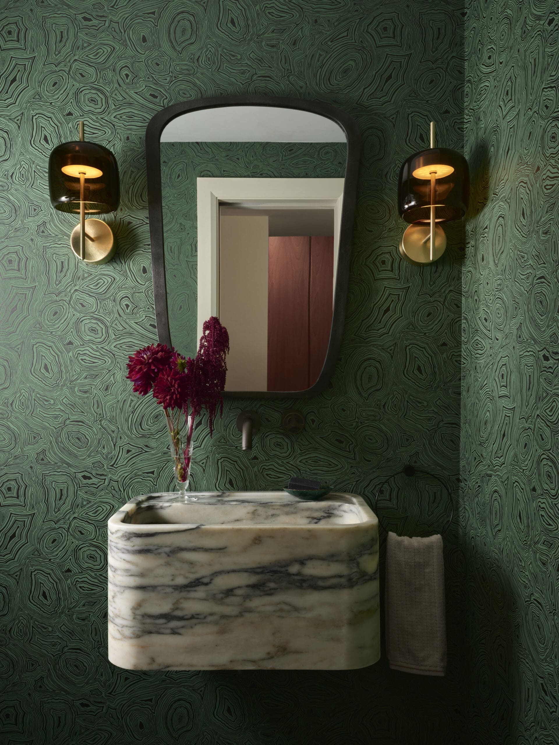 Green patterned bathroom wall with a tall, rounded-framed mirror above a marble pedestal sink and two brass sconces with amber glass shades.