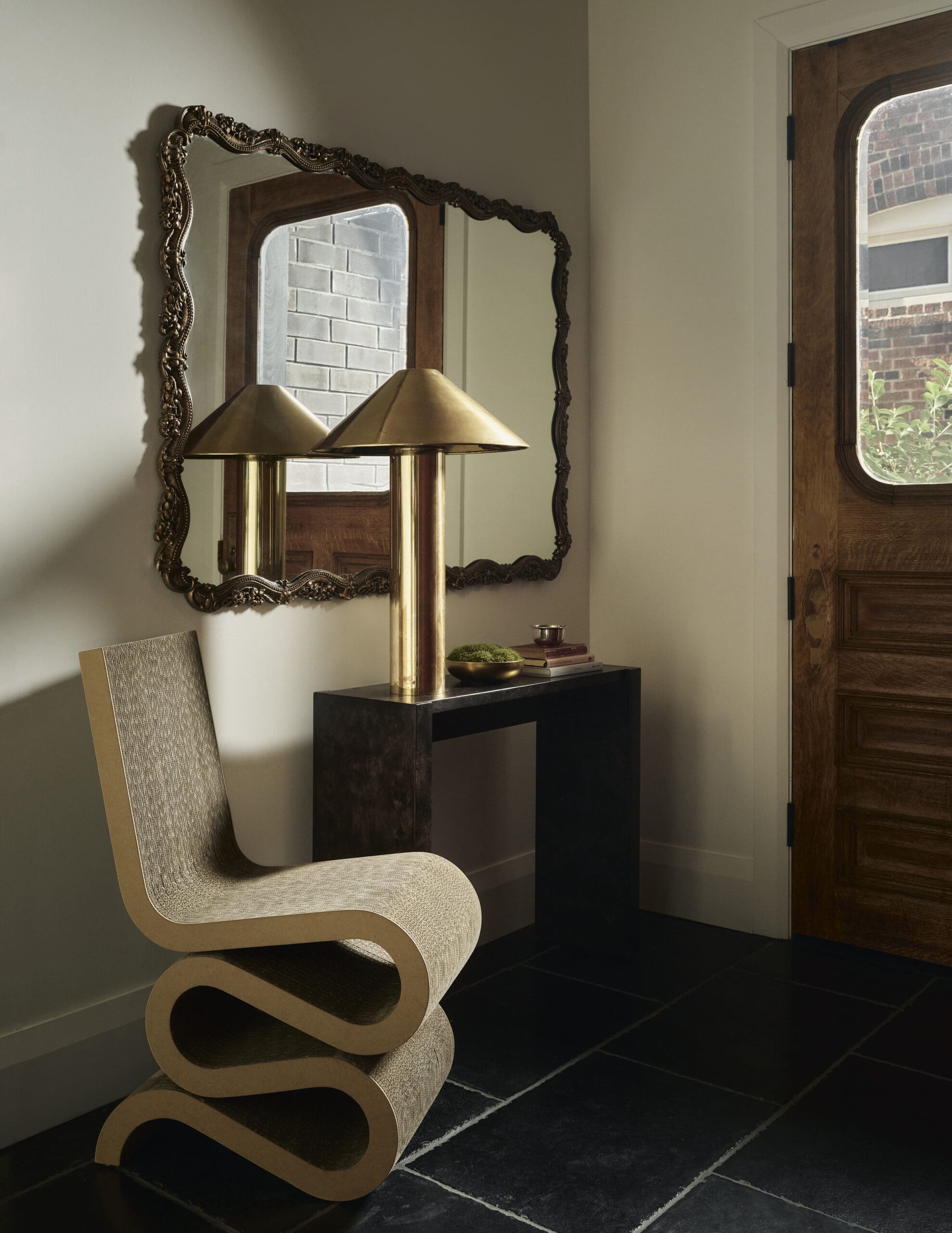 Interior entryway with a sculptural curved beige chair, a gold cylindrical lamp, and an ornate framed mirror above a dark console. A wooden door with glass window is to the right.