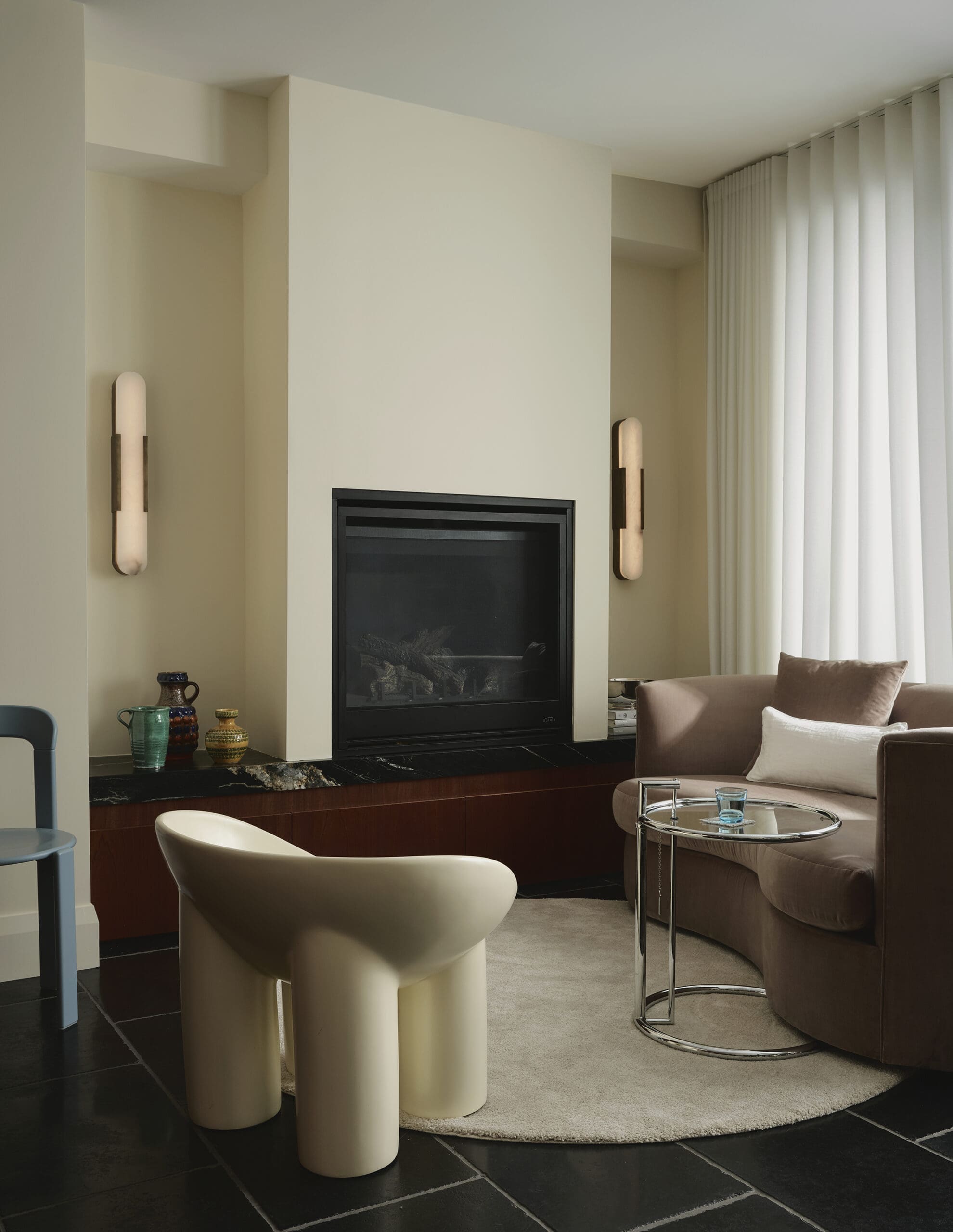 Contemporary living room with a centered fireplace, beige walls, and sculptural ivory stools beside a round glass table.