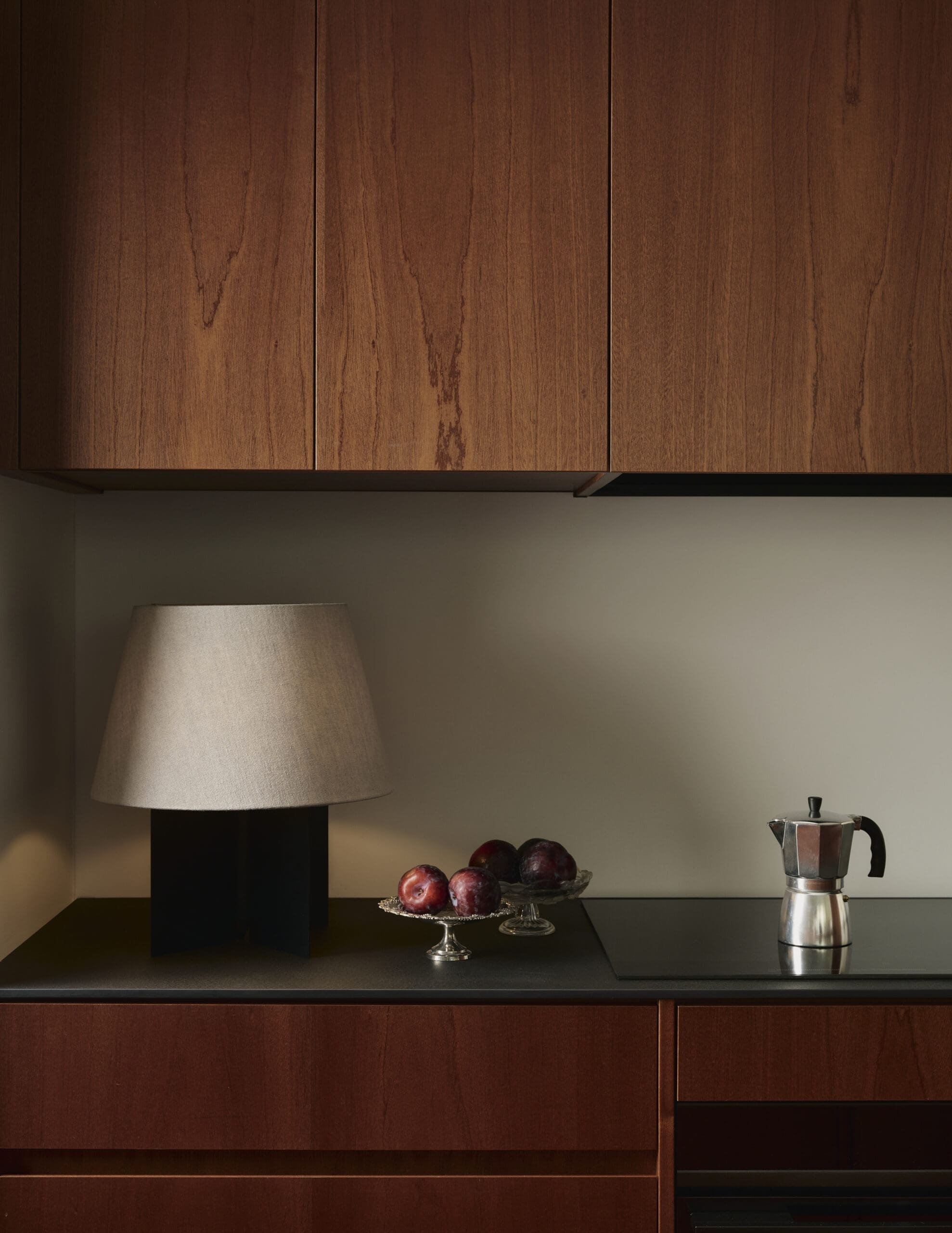 Wooden kitchen cabinets above a dark countertop, with a lamp on the left and a bowl of apples paired with a moka pot on the right.