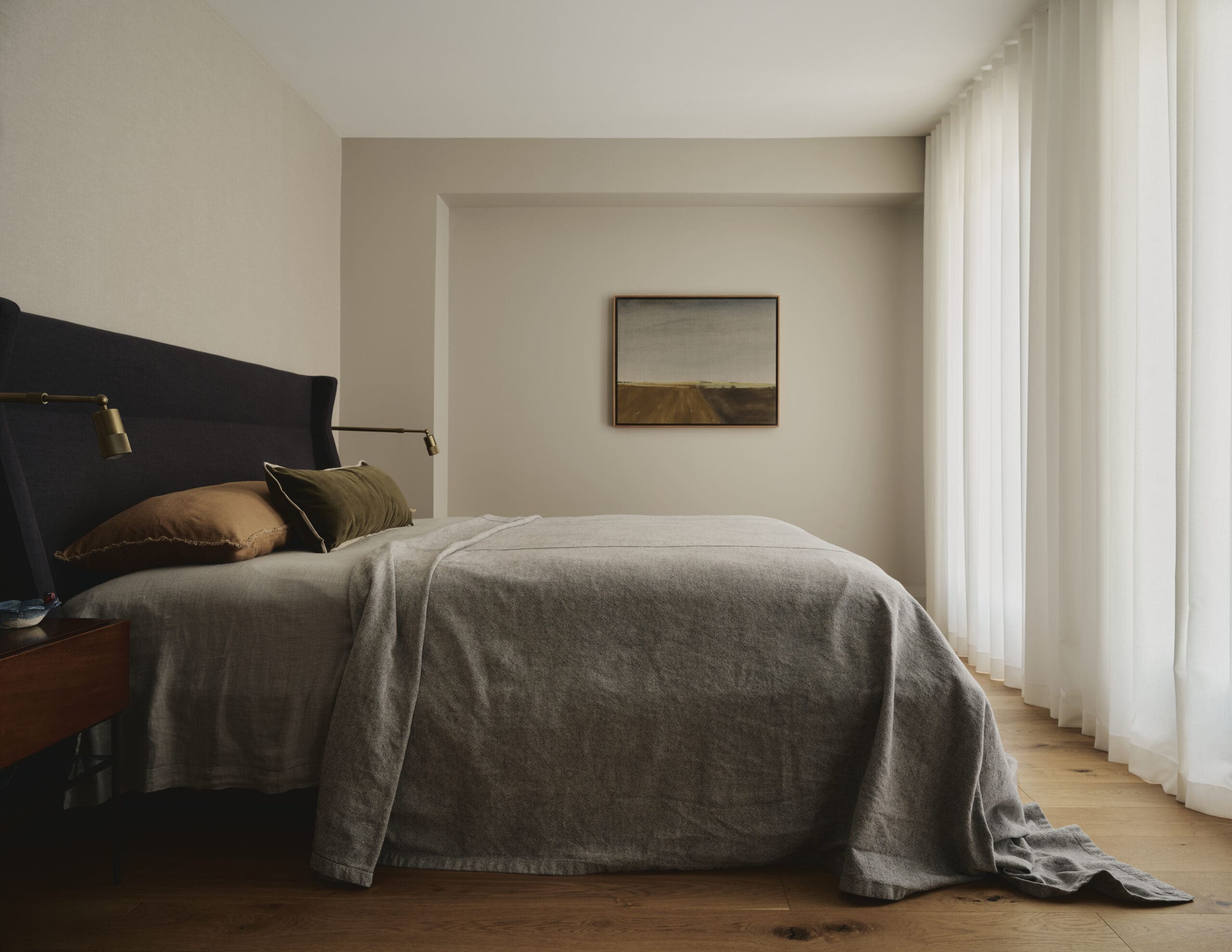 A neatly made bed with a dark headboard, gray bedding, and brown pillows in a softly lit bedroom with sheer white curtains along the right wall.