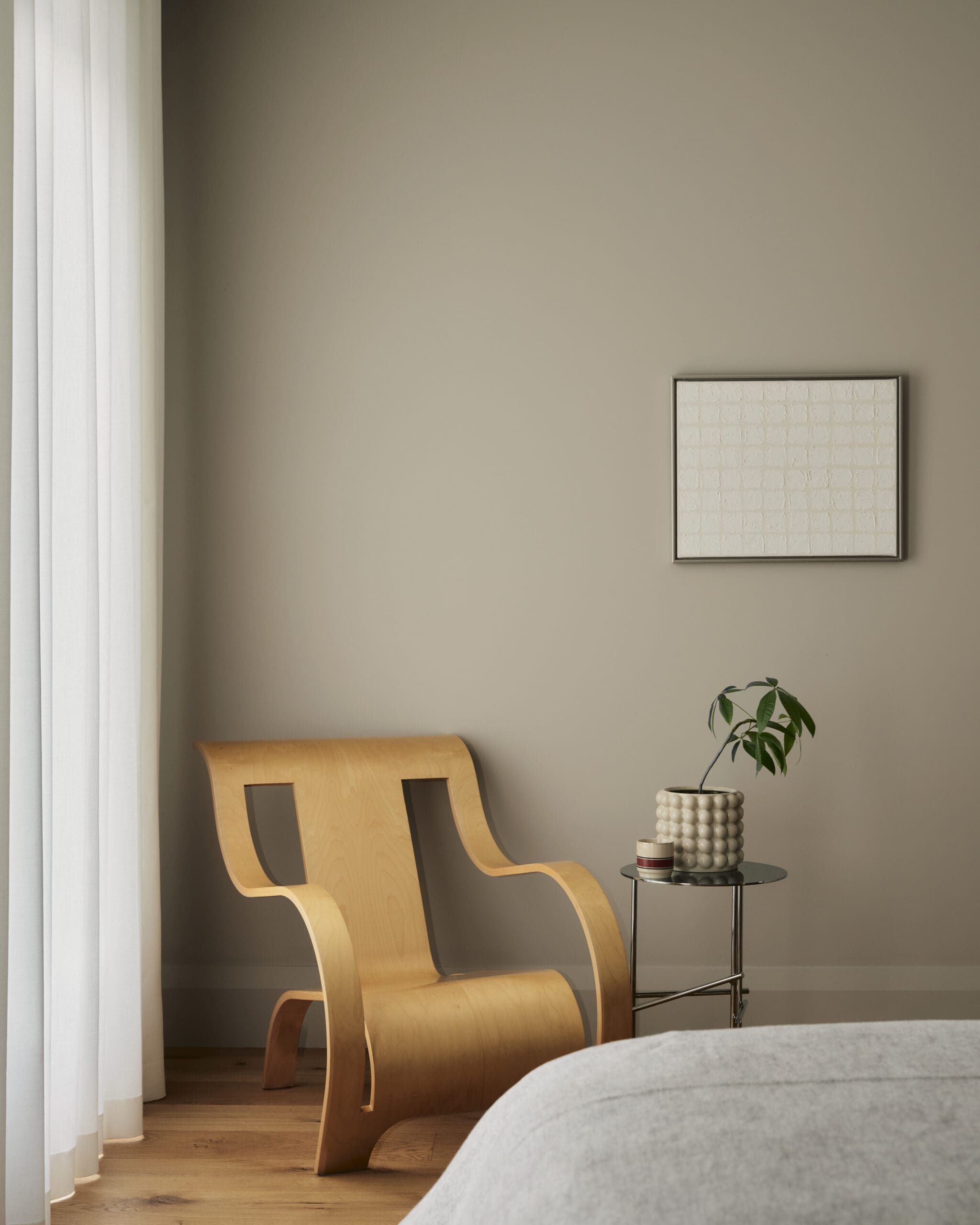 Corner of a living room with two curved wooden chairs, a small round table with a potted plant, beige wall, and a framed textured square artwork on the wall.