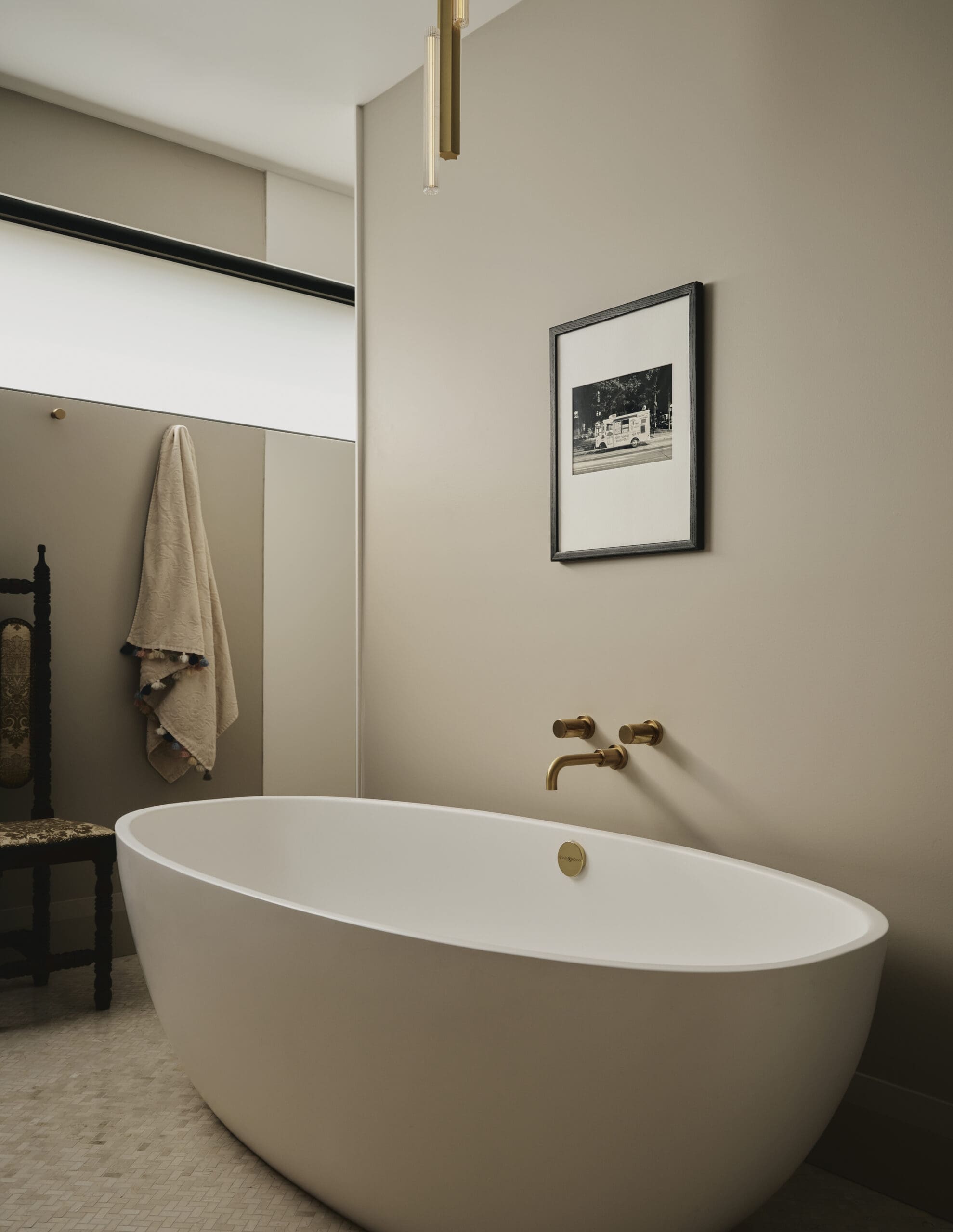 Modern bathroom with a white freestanding bathtub, gold faucet fixtures, beige walls, and a framed black-and-white photo on the wall.
