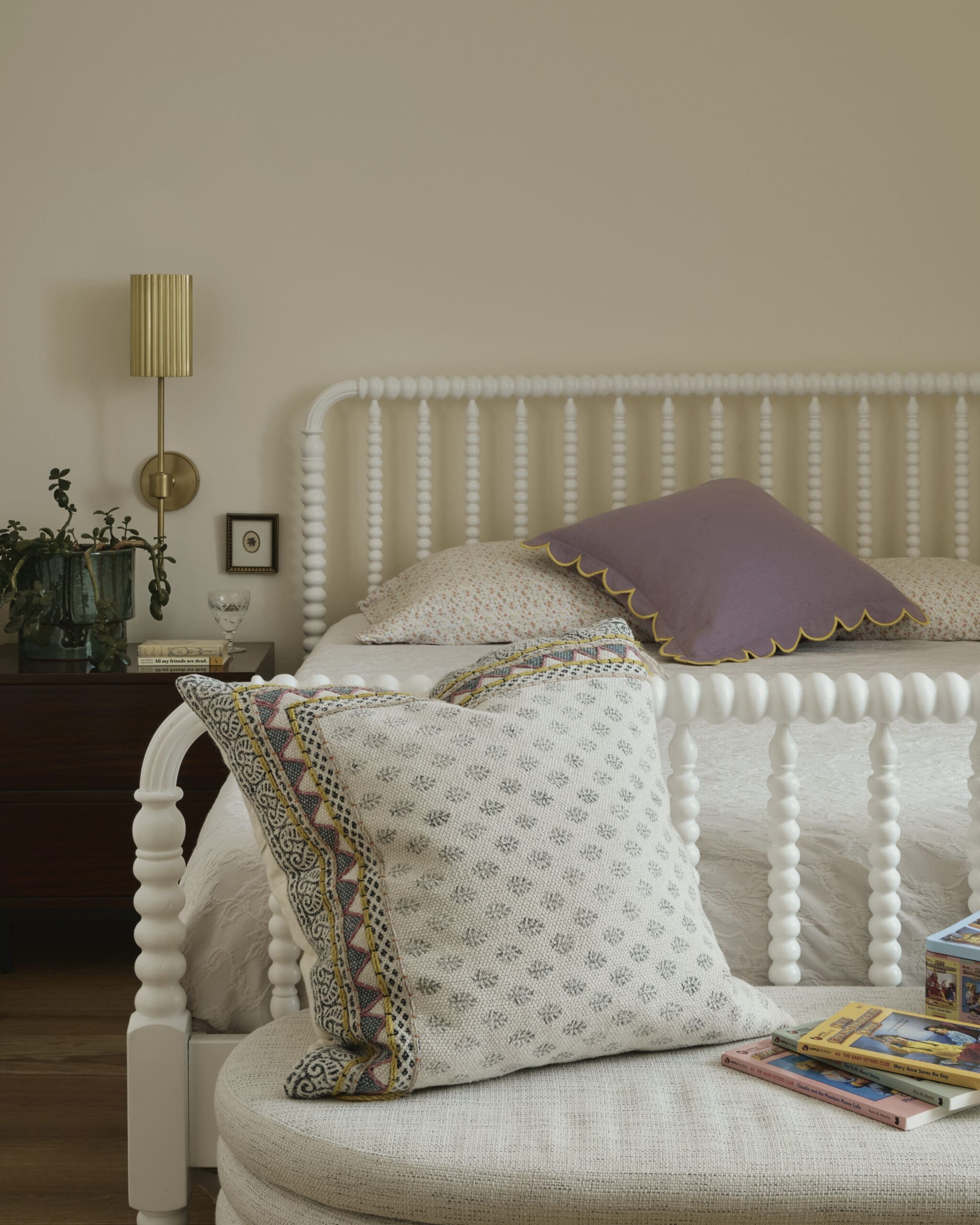 Cozy bedroom corner with a white spindle bed, patterned pillows, and a wooden nightstand with books and plants.