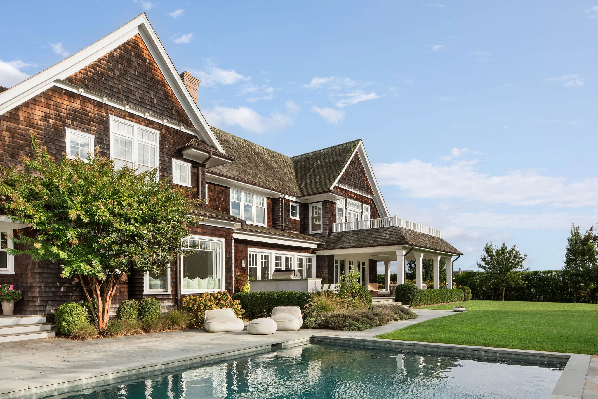Large two-story house with dark wood shingles, white trim, and steep gabled roofs, overlooking a rectangular reflecting pool and manicured lawn under a blue sky.