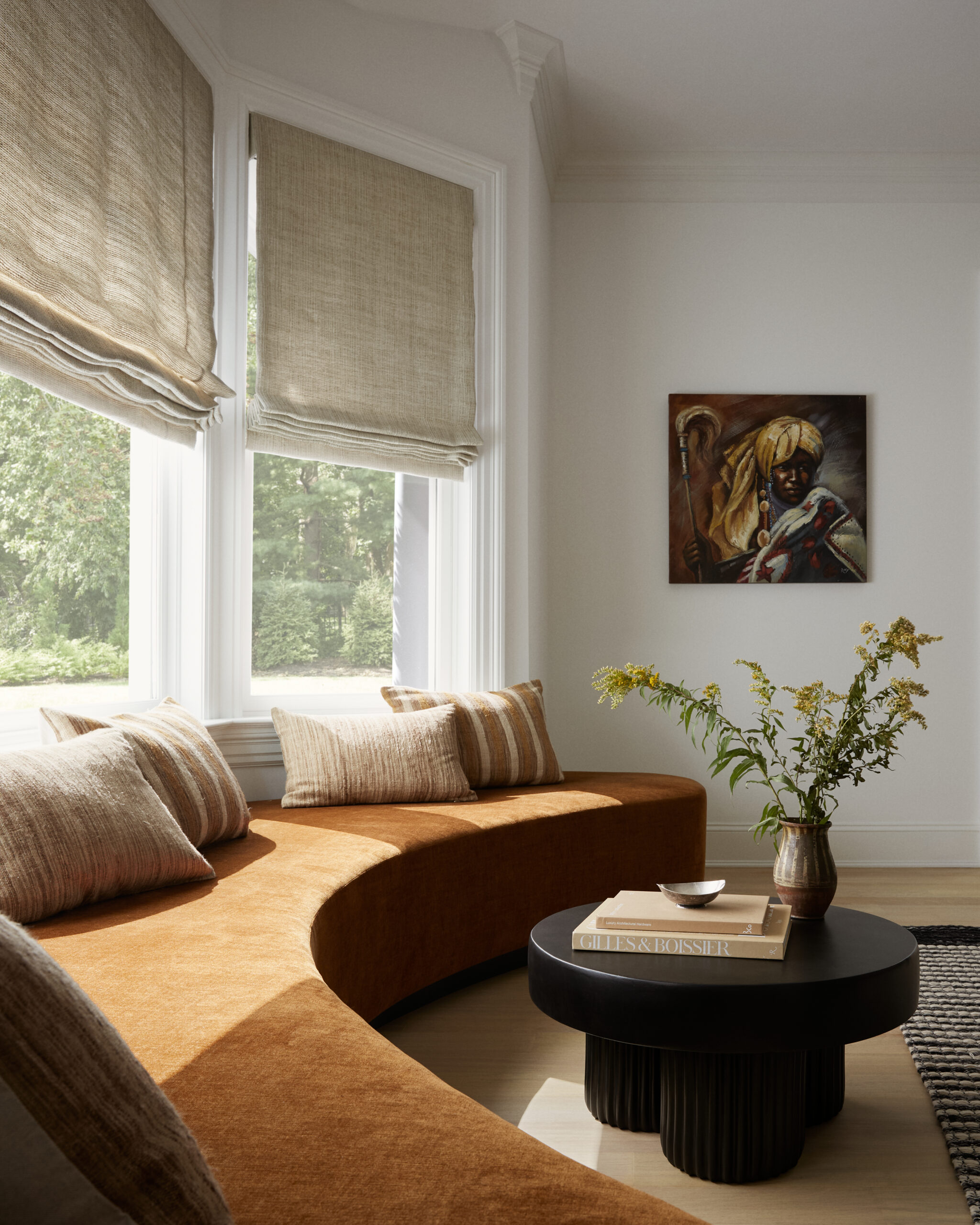 Bright living room with an orange curved sofa, beige striped cushions, large windows with linen blinds, a round black coffee table, and a portrait painting on the wall.
