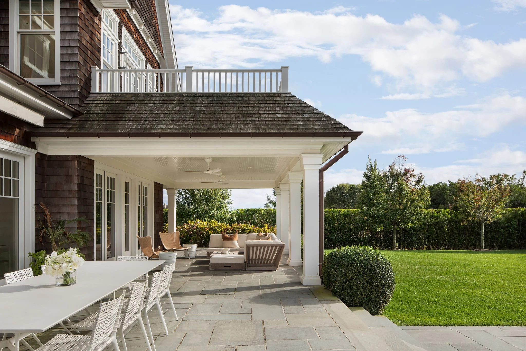 Backyard patio of a house with a long white dining table, wicker chairs, and a seating area under a covered veranda.