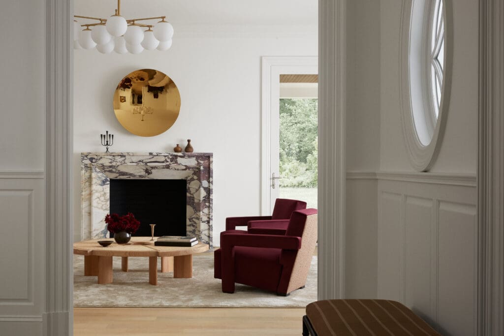 Living room with a marble fireplace, a large round gold mirror, and a modern brass chandelier, flanked by red upholstered chairs.