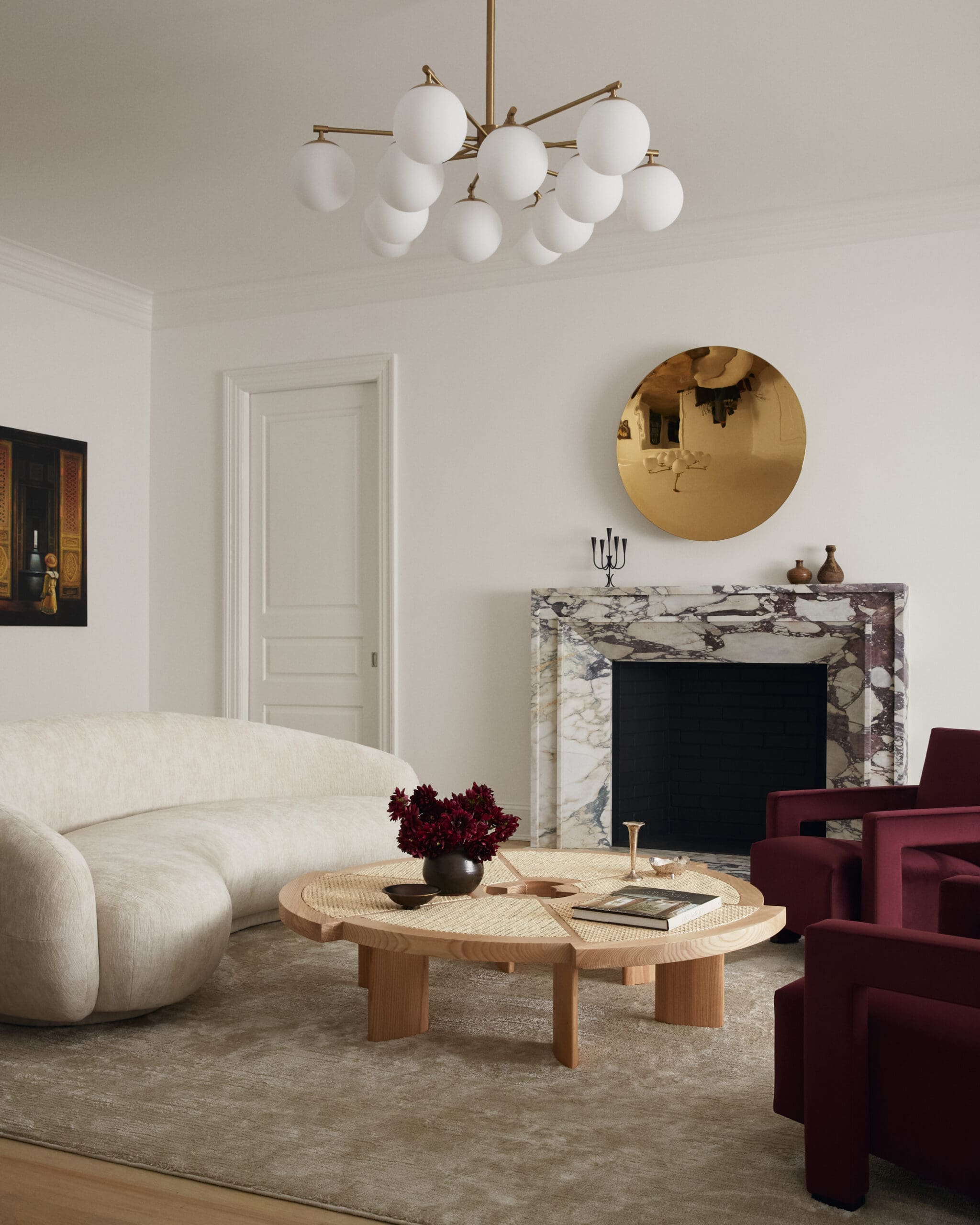 Beige living room with a marble fireplace, round gold mirror above, and burgundy chairs around a wooden coffee table.
