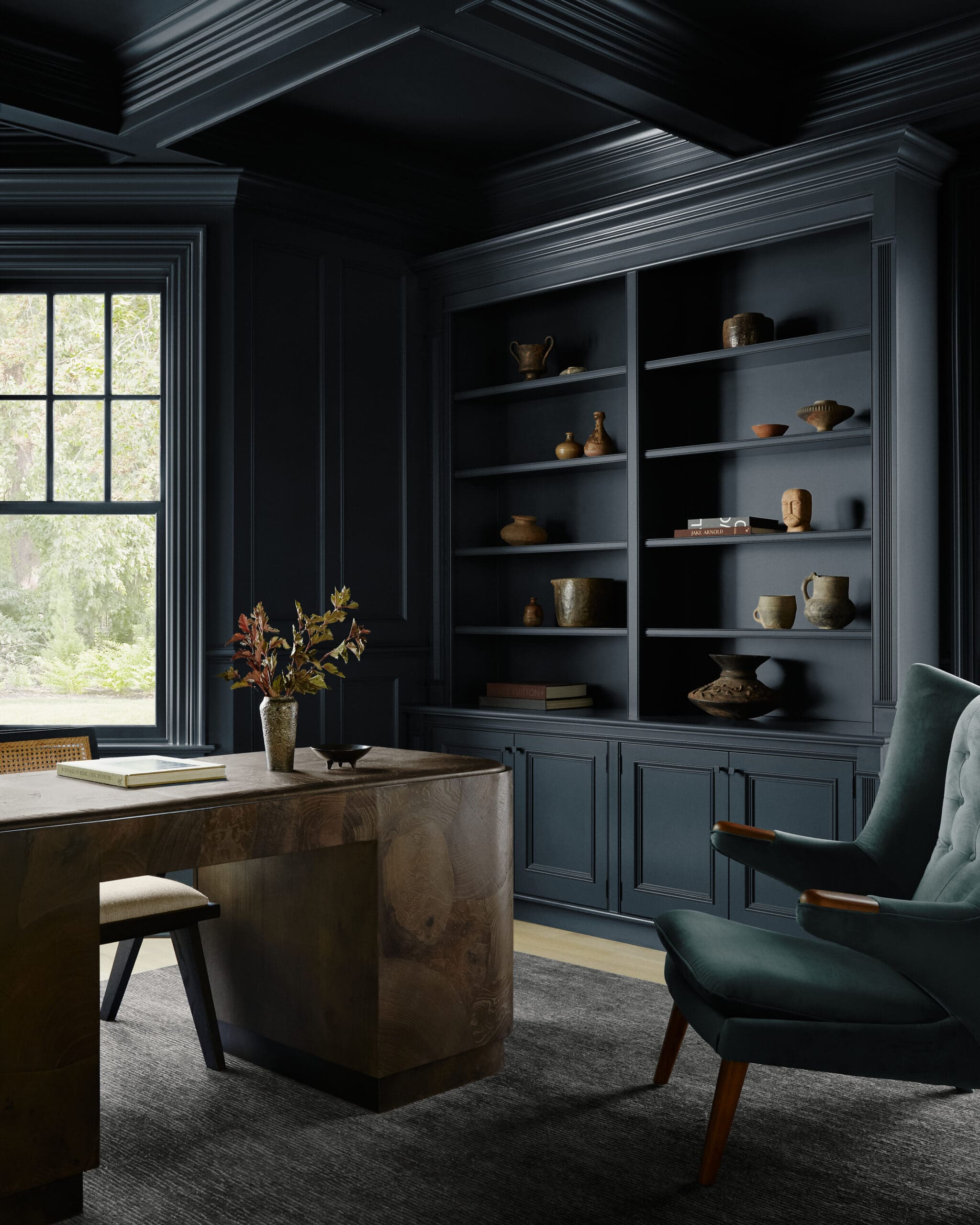 Navy built-in bookshelf with multiple shelves displaying ceramic pots and bowls, next to a window in a dark, cozy room.
