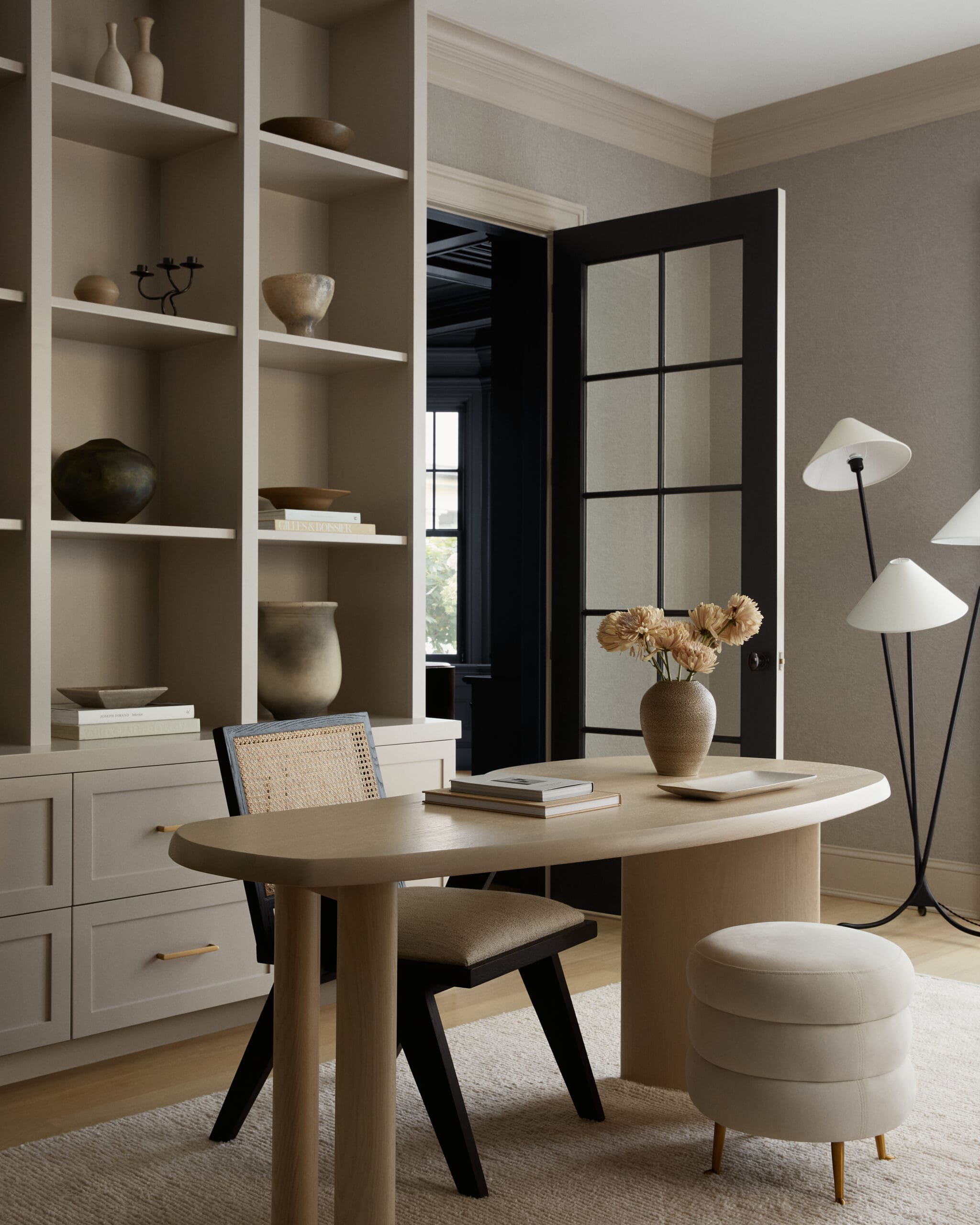 Living room workspace with tall beige shelving filled with pottery, a round wooden table, and a glass-paneled door in the background.