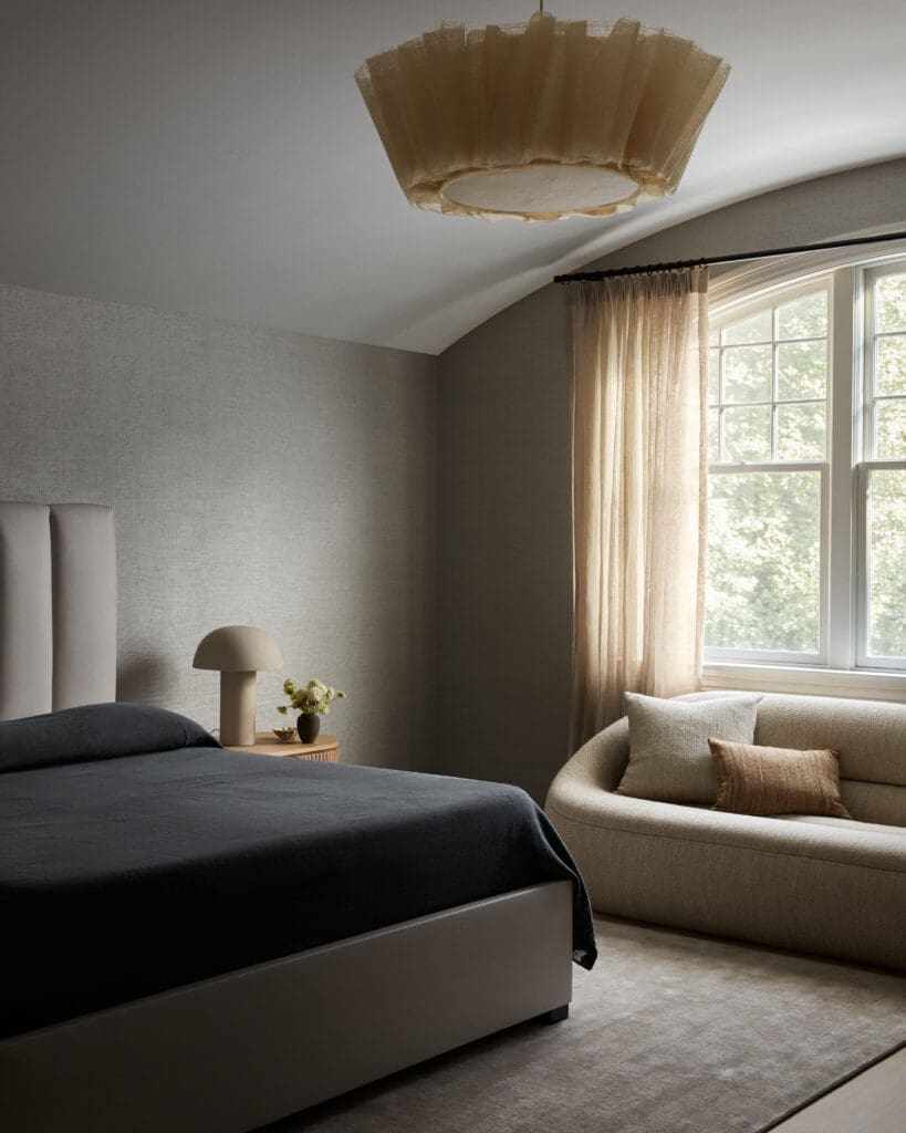 Minimalist bedroom with a low bed, dark bedding, and beige lampshade on a light wood nightstand by a window with sheer curtains.