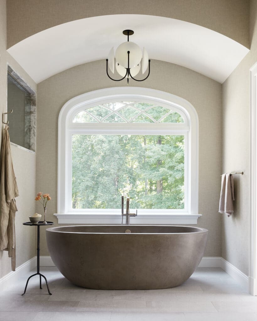 Freestanding oval bathtub under an arched window with a black chandelier above in a beige-toned bathroom.