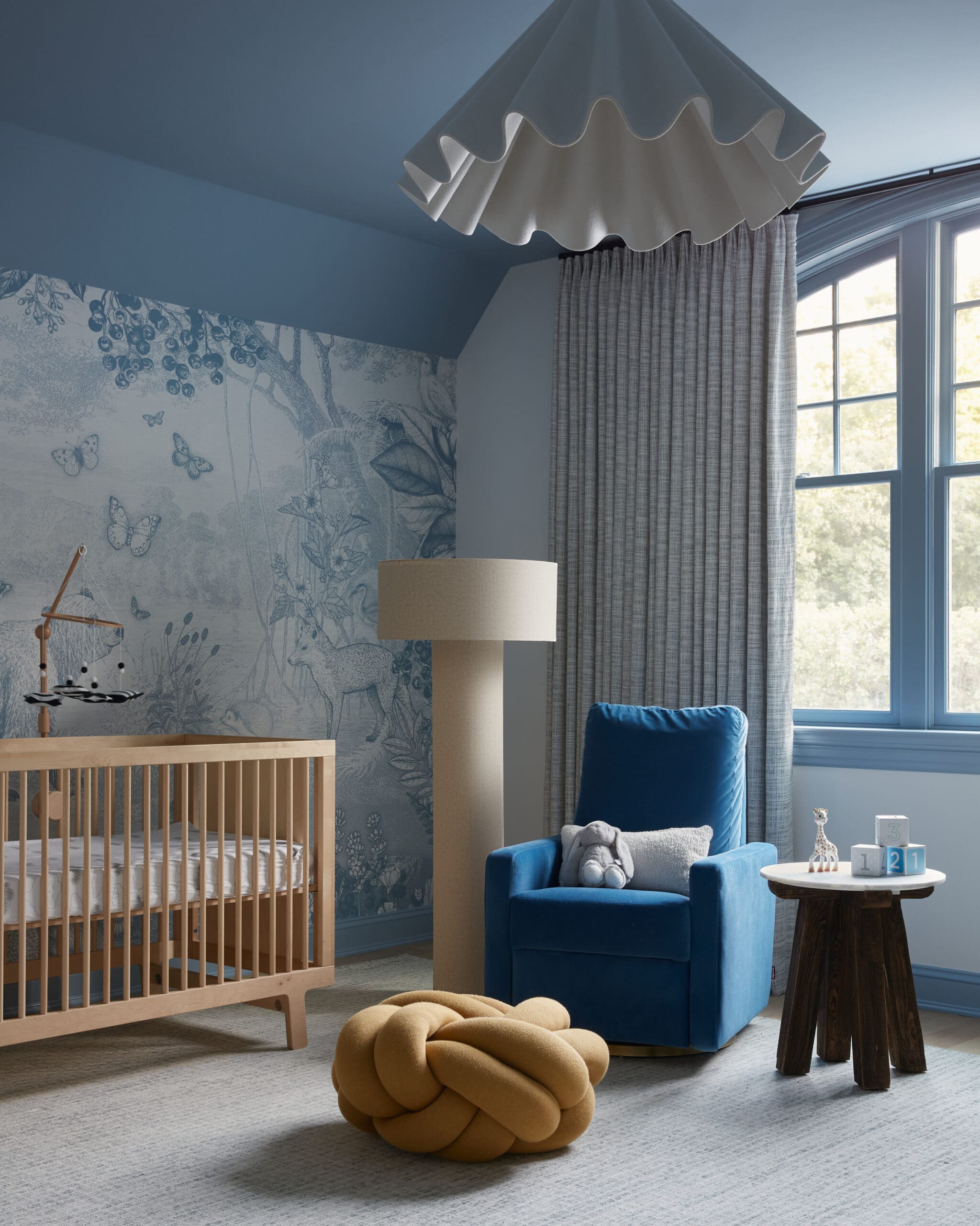 Blue-toned nursery with a wooden crib, blue chair, patterned wallpaper, and large window with gray curtains.
