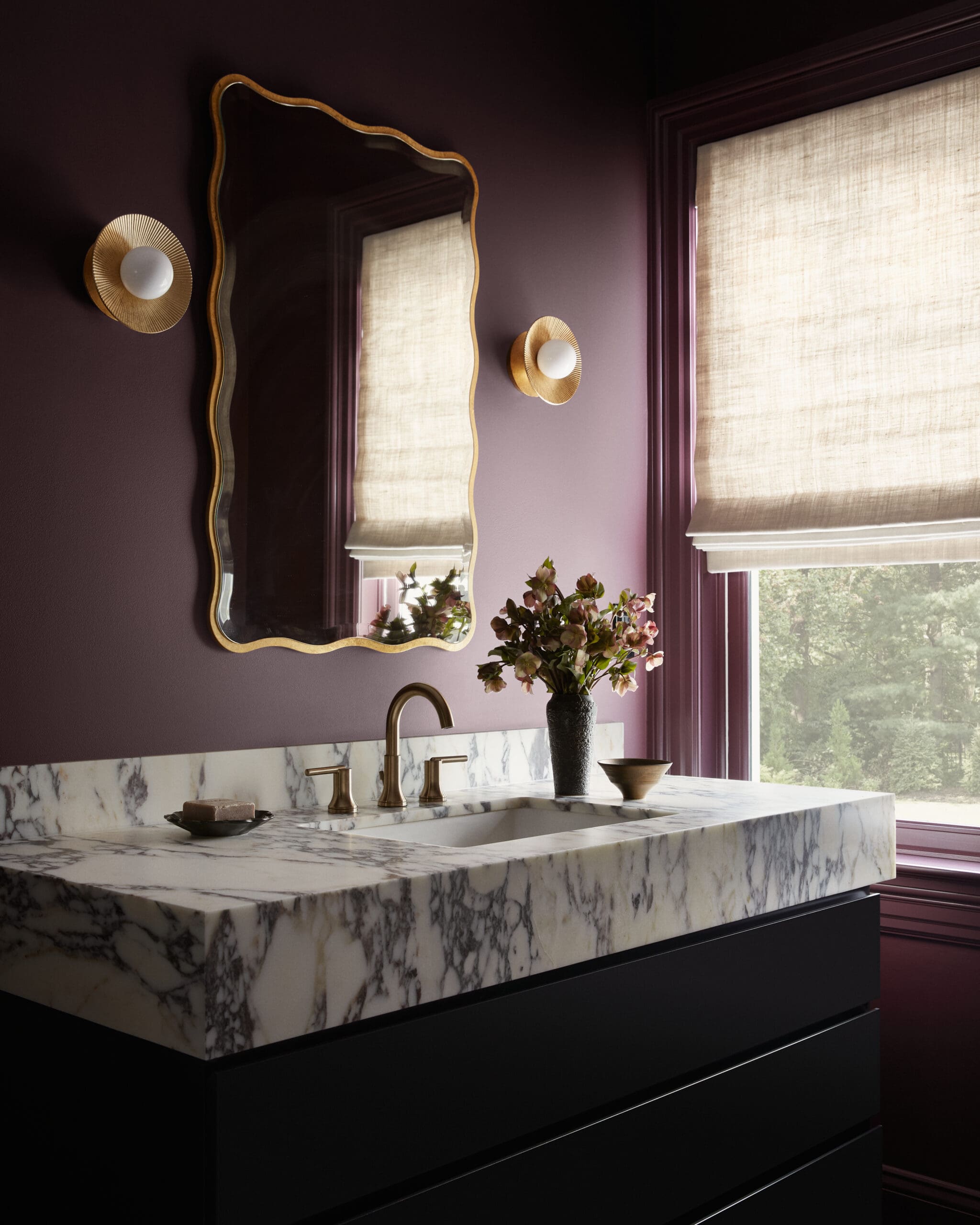 Purple-walled bathroom with a marble countertop vanity, brass faucet, and a wave-edged gold-framed mirror flanked by round gold wall sconces.