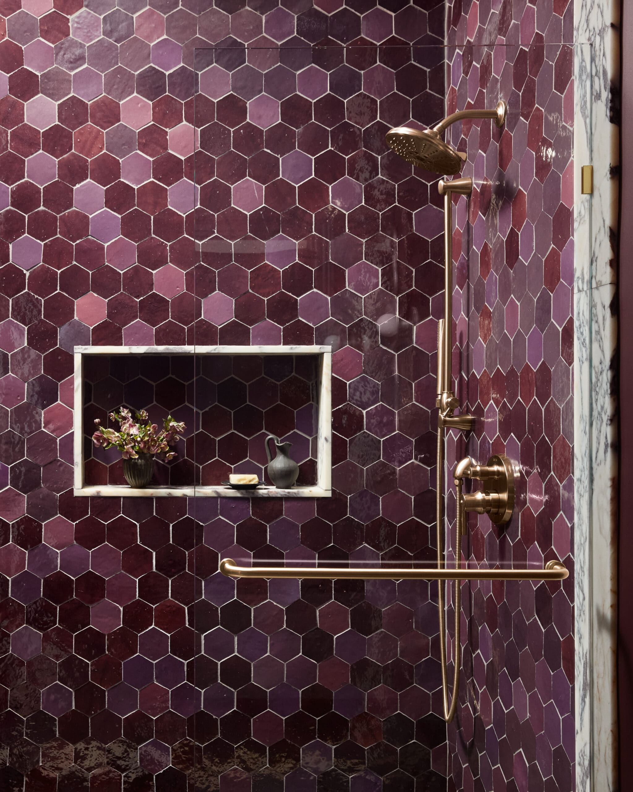 Bronze shower fixtures mounted on a burgundy hex tile wall with a marble-framed recessed alcove holding plants and small items.