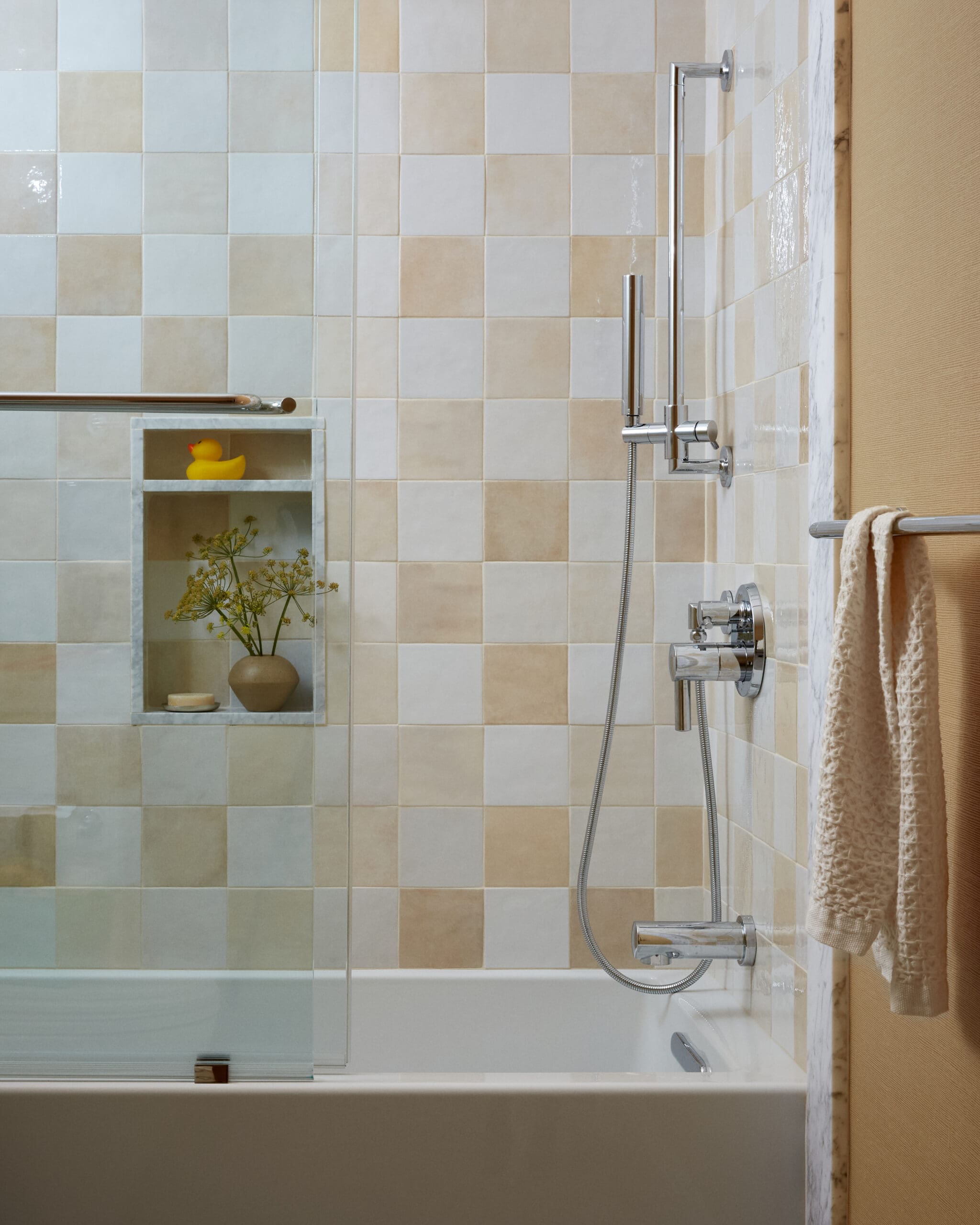 Shower area with beige and light blue checkered tiles, glass door, and chrome handheld shower.