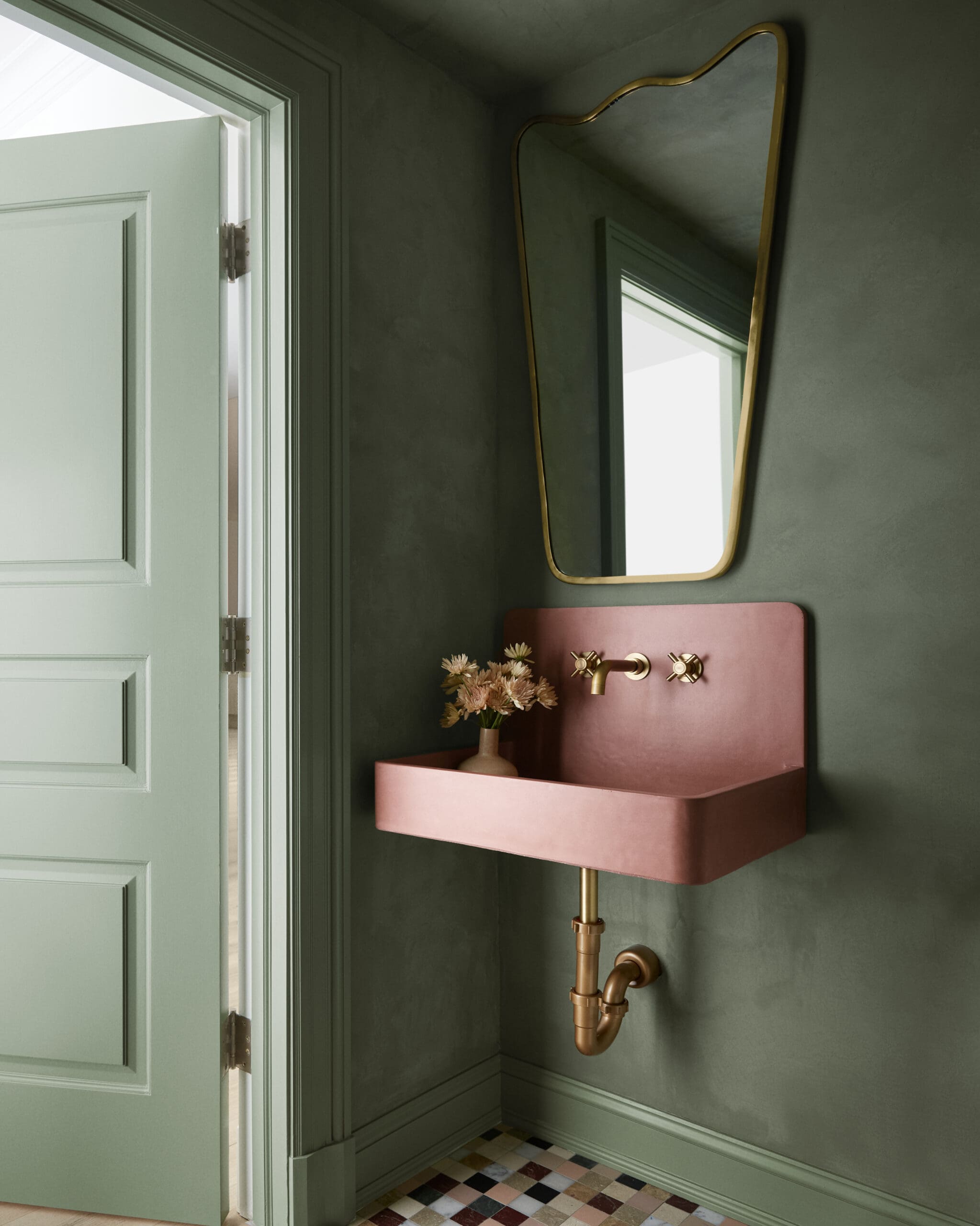 Mint-green door and dark-green wall with a pink copper sink and brass fixtures beneath a gold-framed mirror.