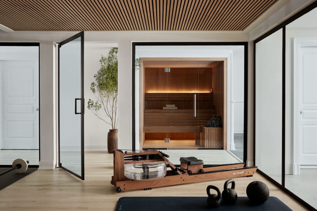 Home gym with a wooden Pilates reformer, kettlebells, and a medicine ball; glass walls reveal a sauna in the background.