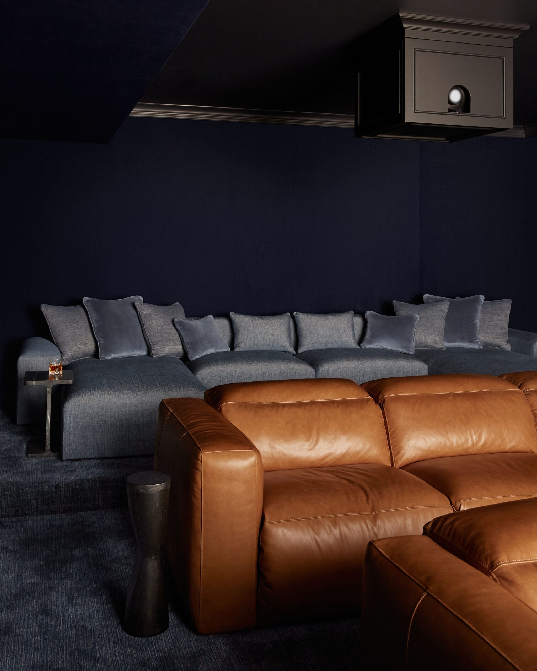 Dark navy living room with a long blue sofa and many gray cushions; brown leather chairs sit in front in a dimly lit space.