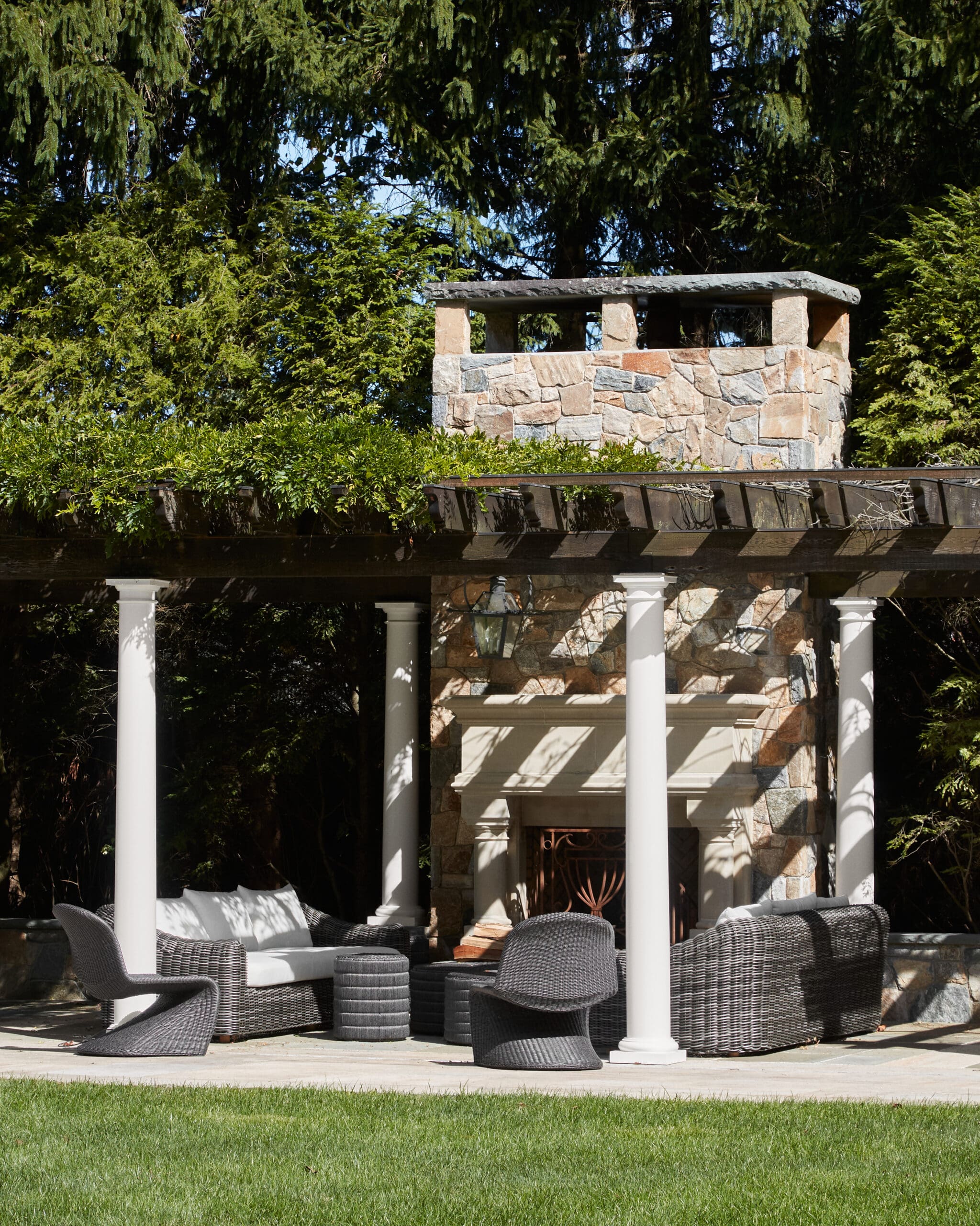 Outdoor patio with wicker seating around a stone wall and columns under a pergola, lush greenery in the background