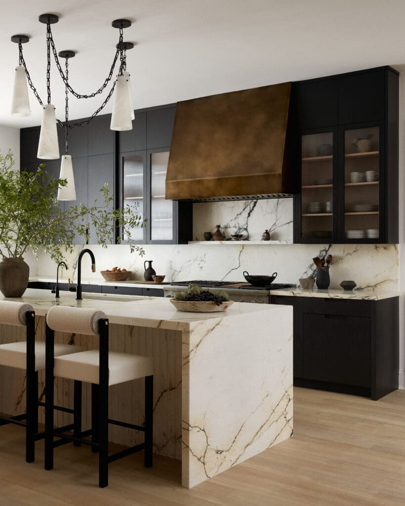 Modern kitchen with black cabinetry, a copper range hood, and marble countertops beneath a chain-hung pendant light fixture.