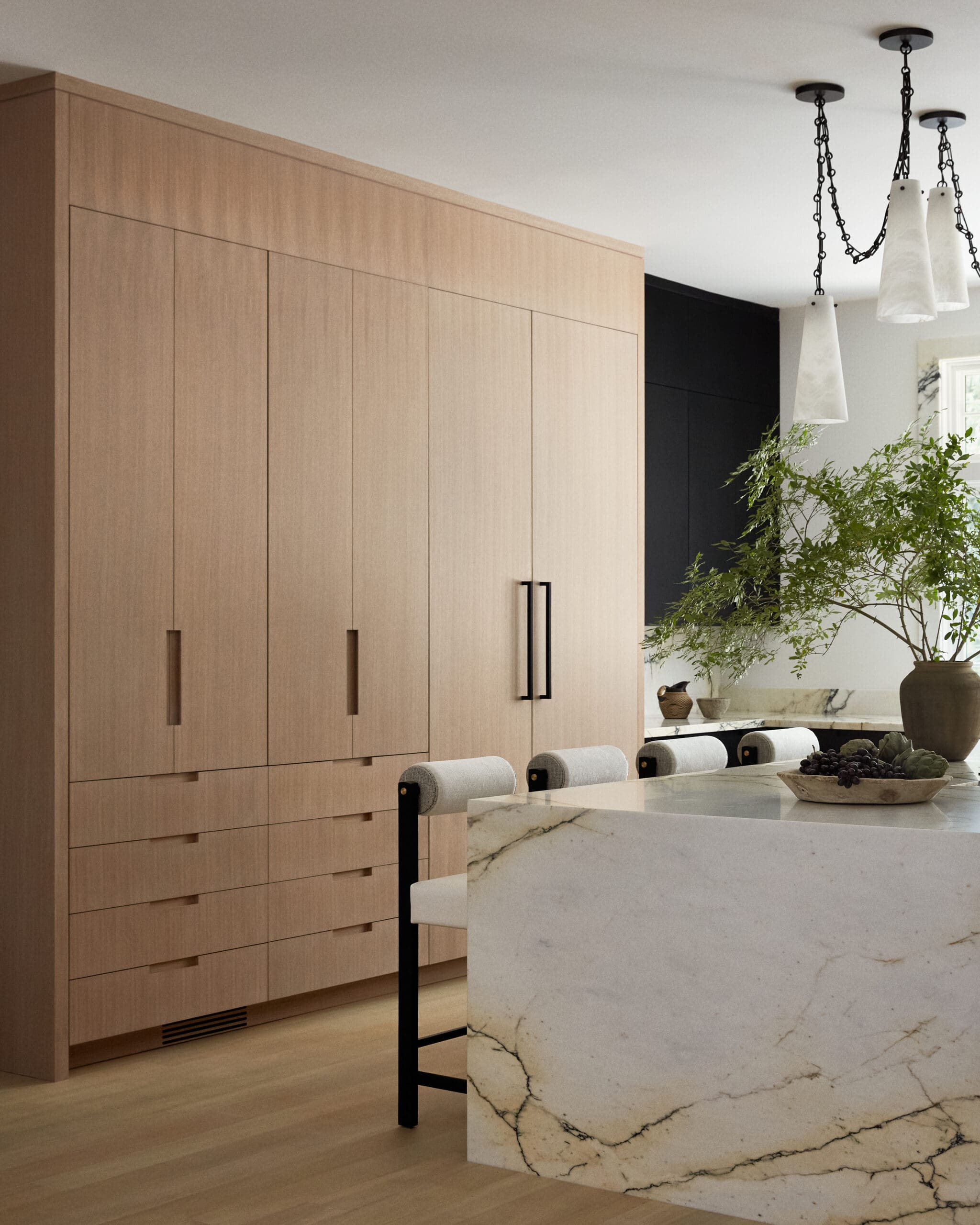 Modern kitchen with tall light wood cabinets along the left wall, and a white marble island with black metal legs in the foreground; large leafy plant visible in the background.