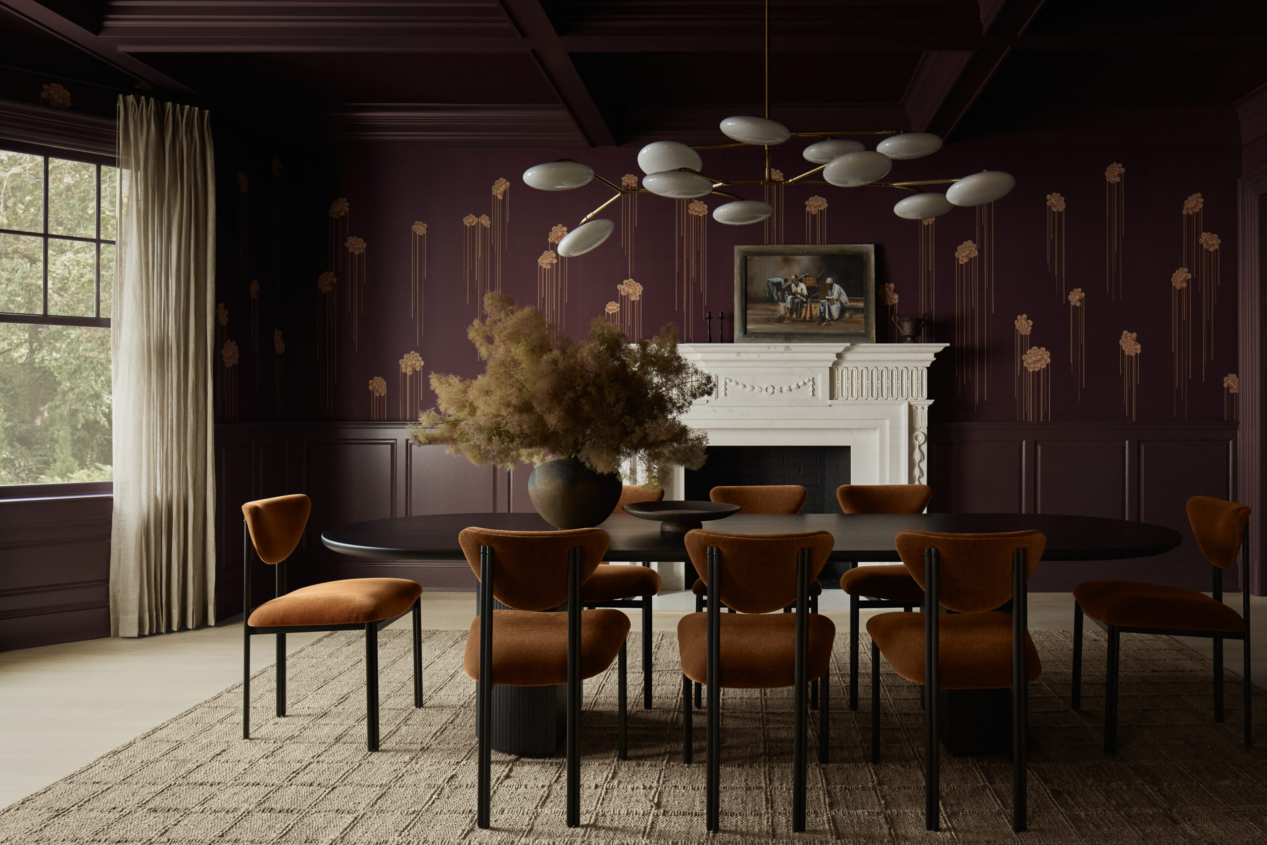 Dark burgundy dining room with a white fireplace mantel, gold wall accents, and a modern chandelier above a round black table surrounded by orange chairs.