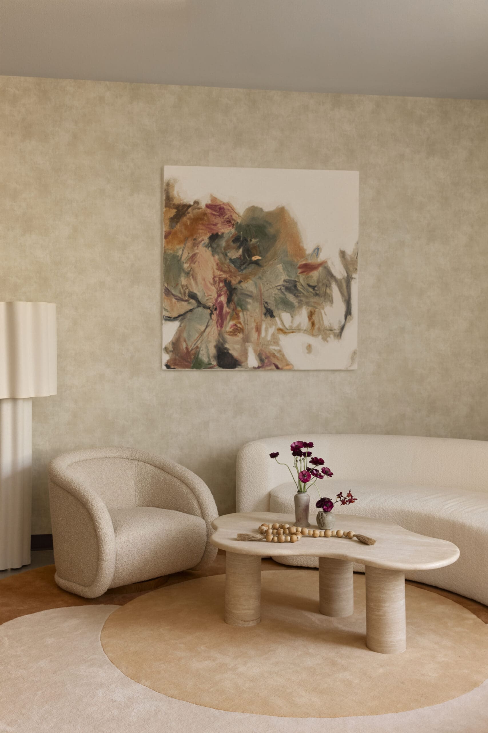 Cozy living room with a beige textured wall, abstract painting, cream seating, and a round coffee table with vases of purple flowers.