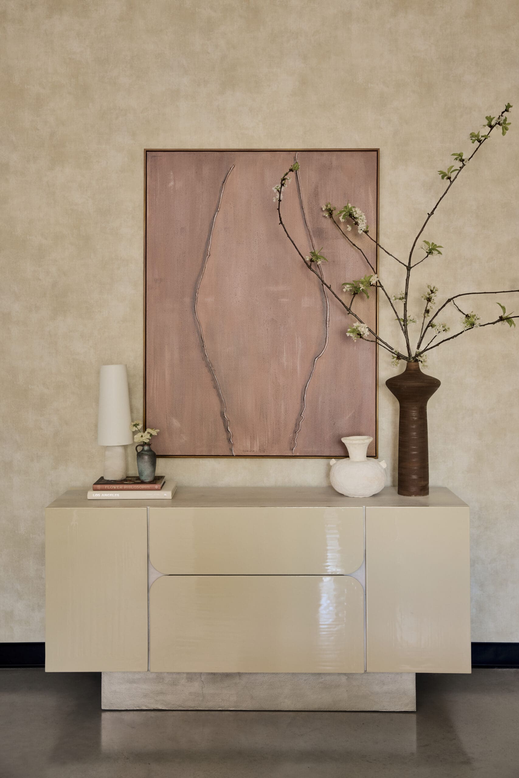Decorative interior scene with a beige glossy cabinet, large pink-framed artwork, a tall white lamp, and a vase with flowering branches on the right.