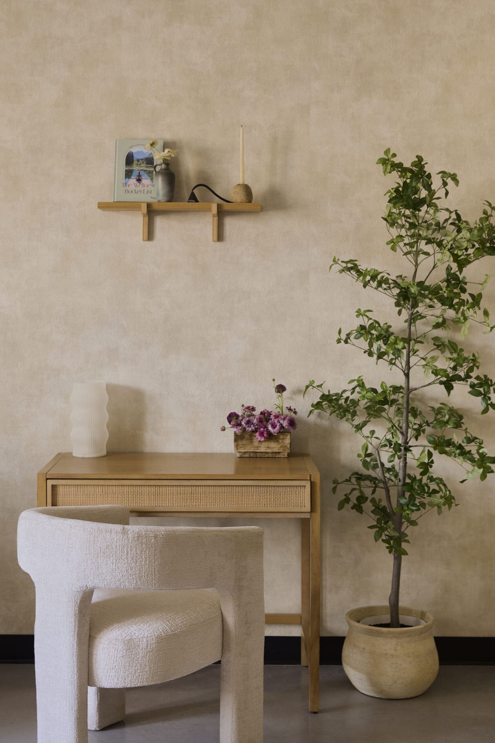 Beige living-room corner with a light wood desk, wall shelf with a book and vase, a candle, and a tall potted plant nearby.
