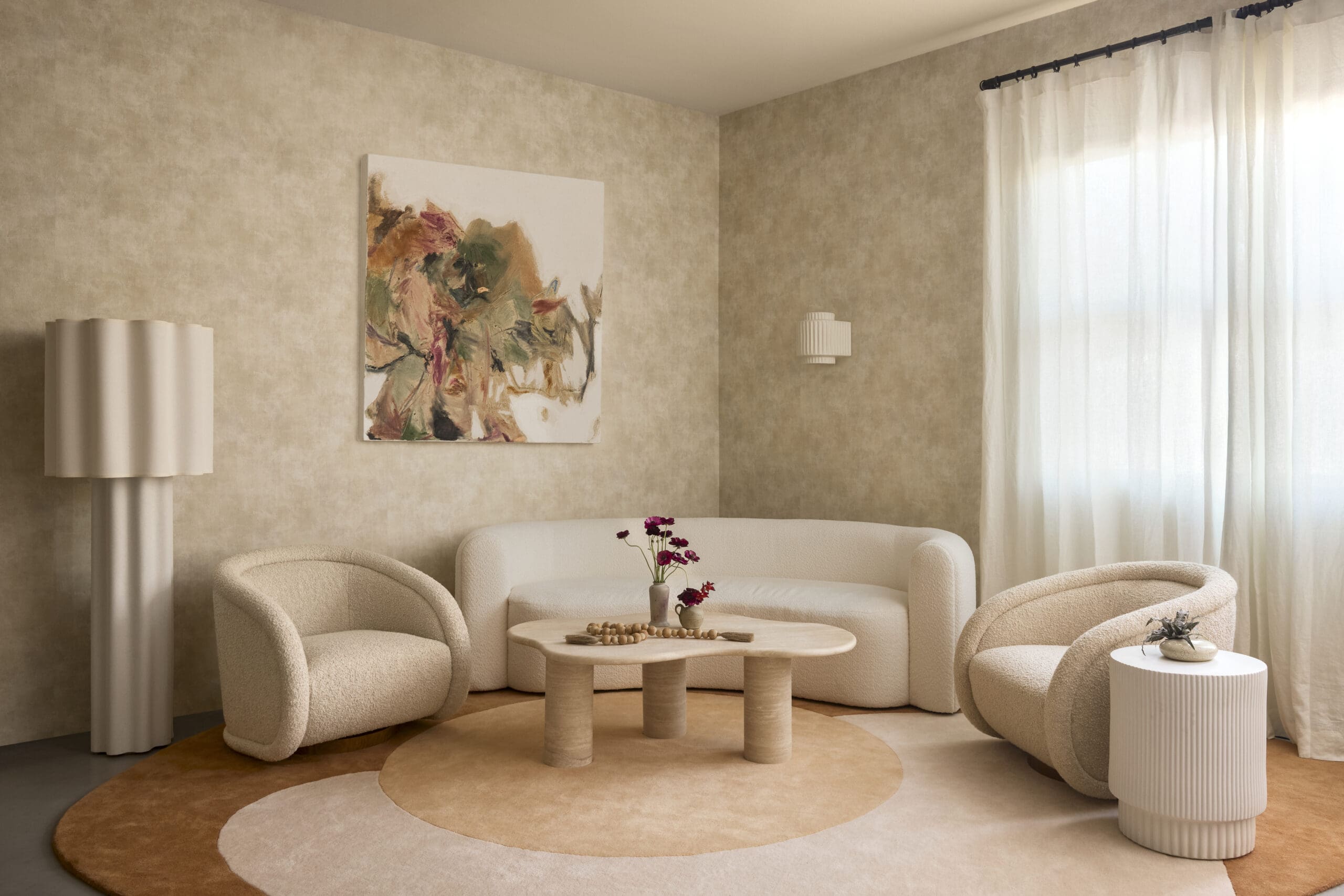 Cozy beige lounge with rounded armchairs, a circular rug, and a light abstract painting on the wall by a white-curtained window