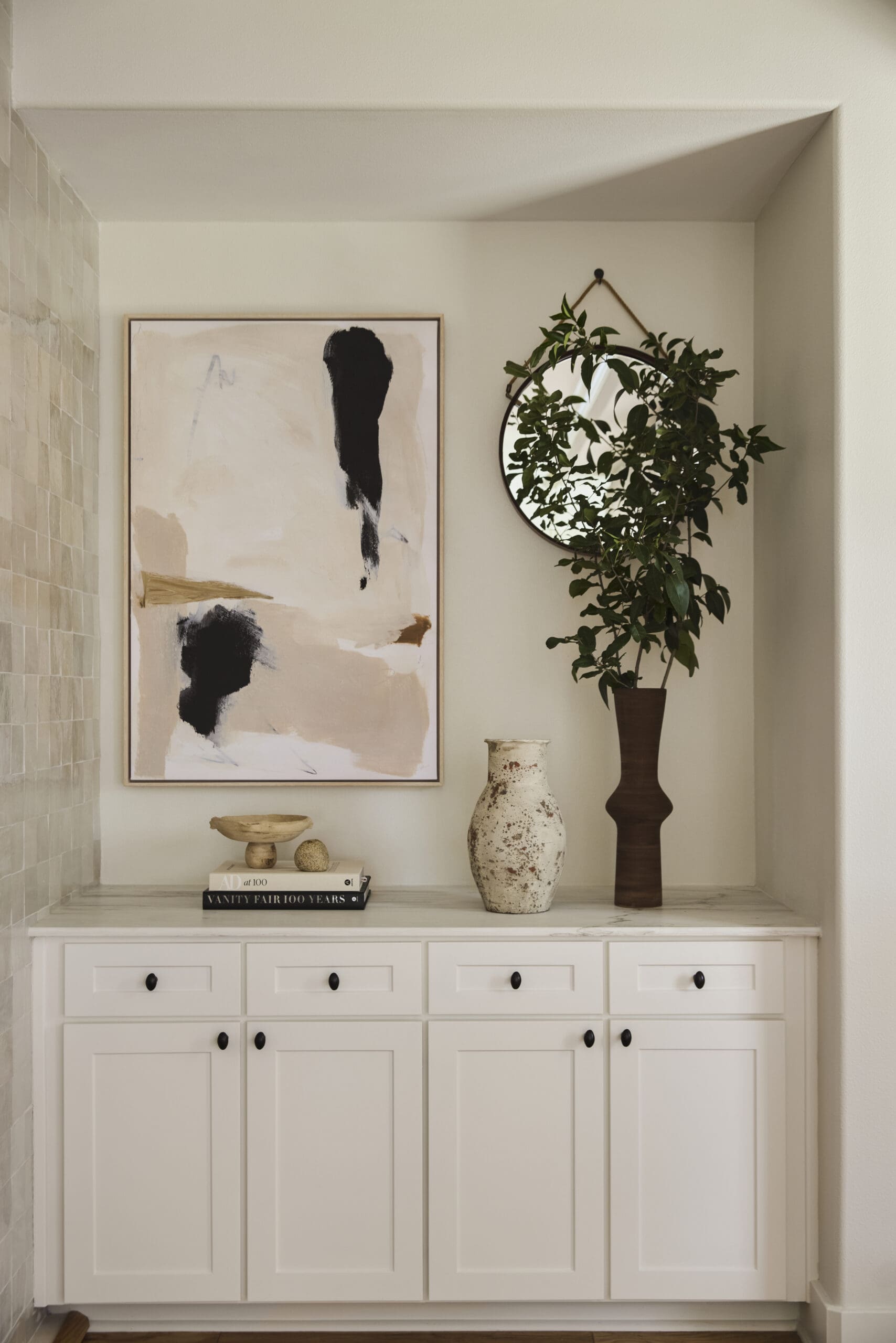 White cabinet with an abstract framed painting, tall leafy plant in a brown planter, round mirror, and ceramic vases arranged on top.