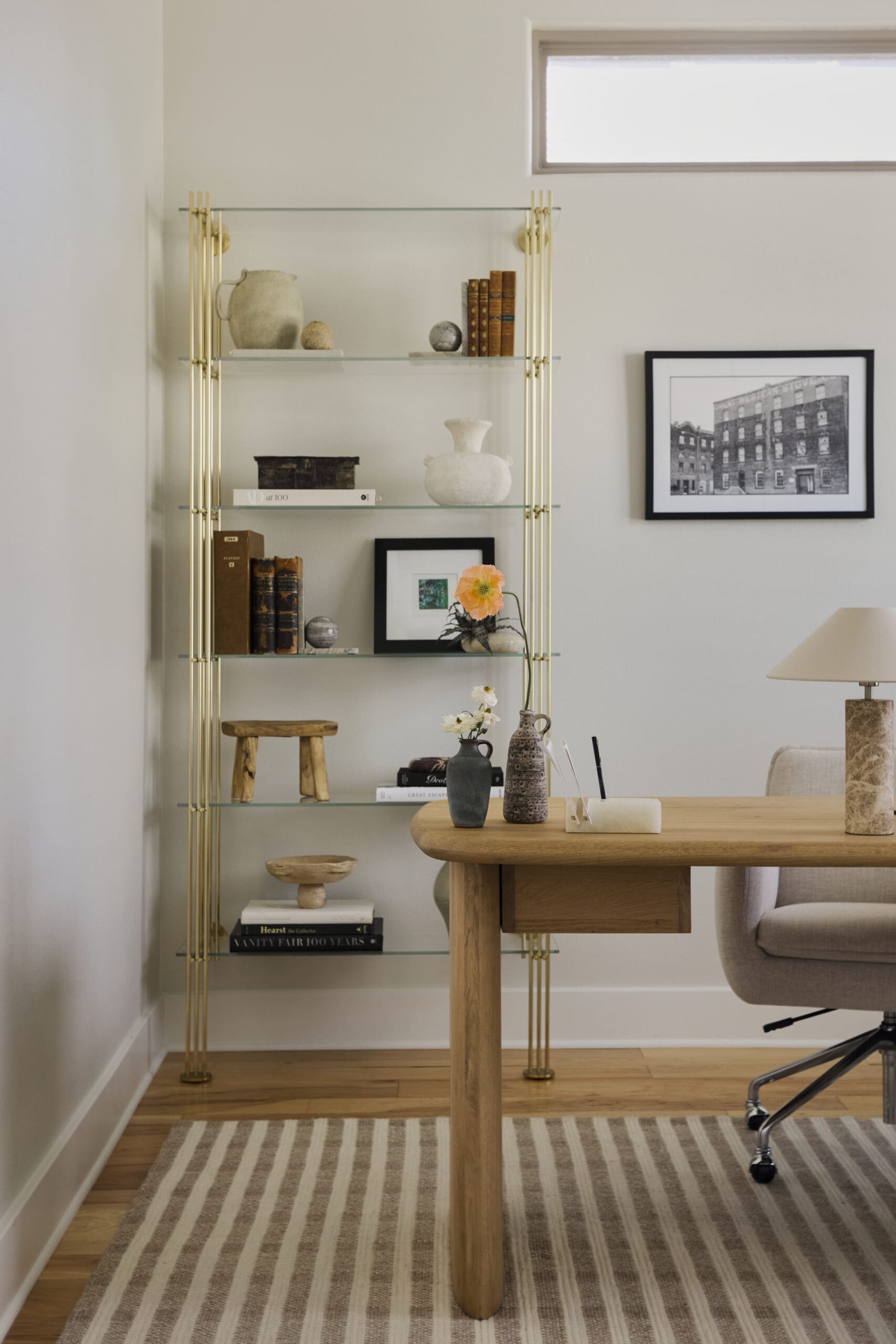 Gold metal-and-glass bookshelf against a pale wall, decorated with pottery, books, and a framed photo on the wall nearby._note: concise alt text shortened for typical readers.