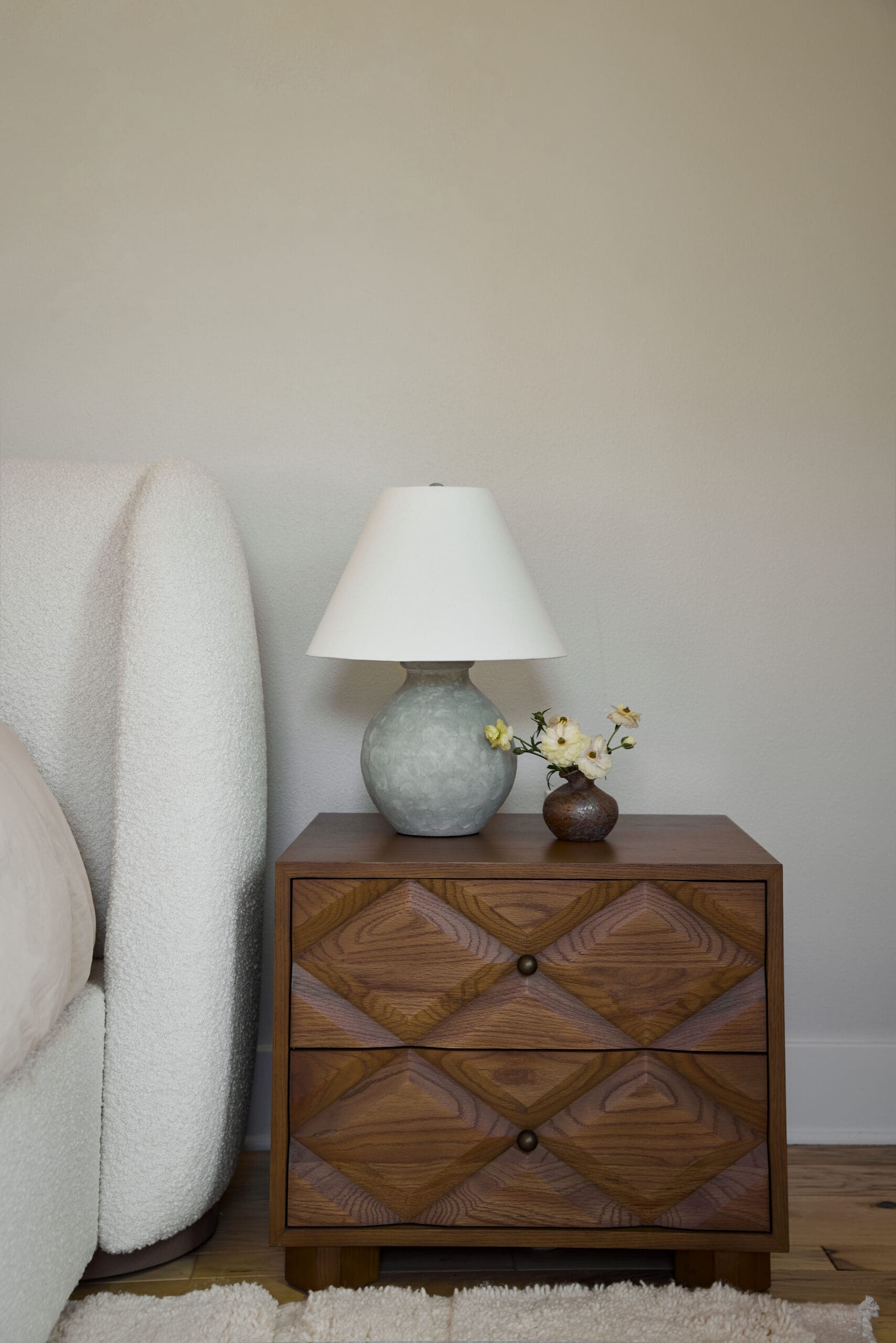 Wooden nightstand with a geometric inlay, holding a pale blue lamp and a small vase of pale yellow flowers, against a beige wall.