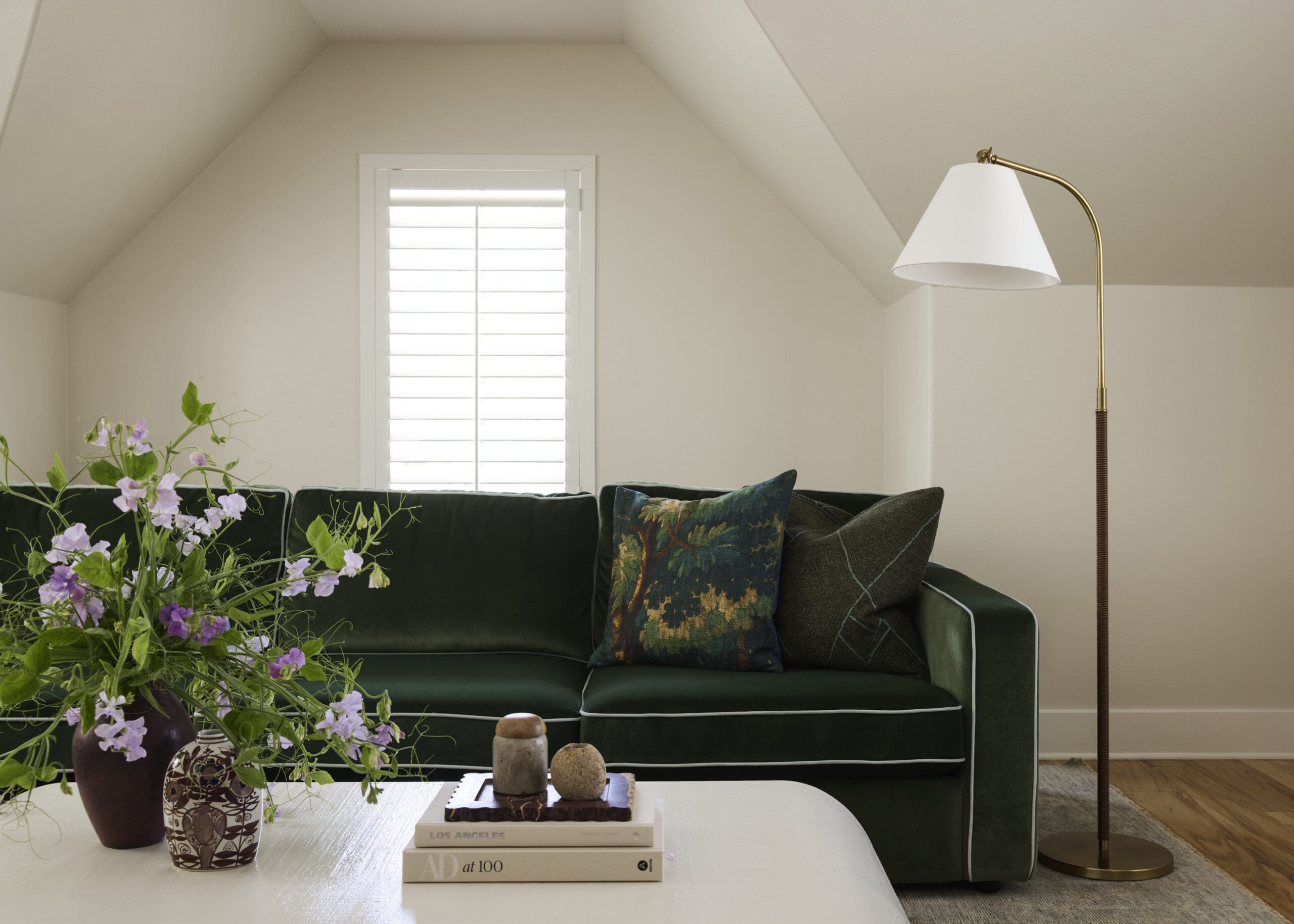 Cozy living room with a dark green velvet sofa, patterned cushions, and a brass floor lamp beside a window with blinds, plus a vase of flowers on a white coffee table.
