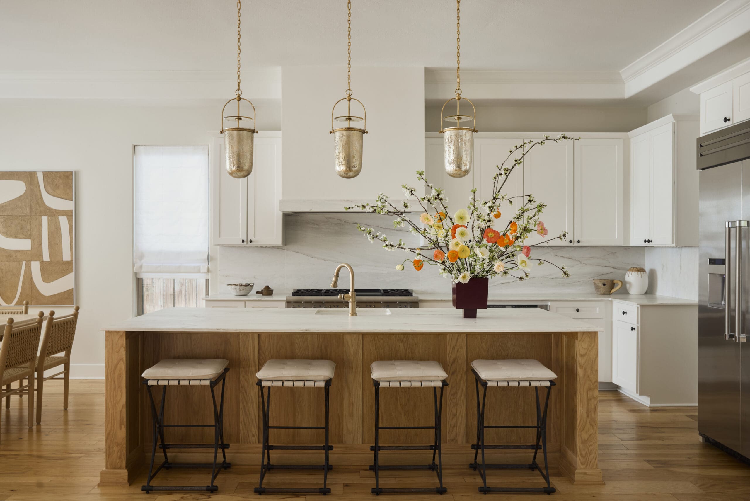 Bright modern kitchen with a wooden island, white cabinetry, and three gold pendant lights over the counter; a vase of colorful flowers sits on the island.
