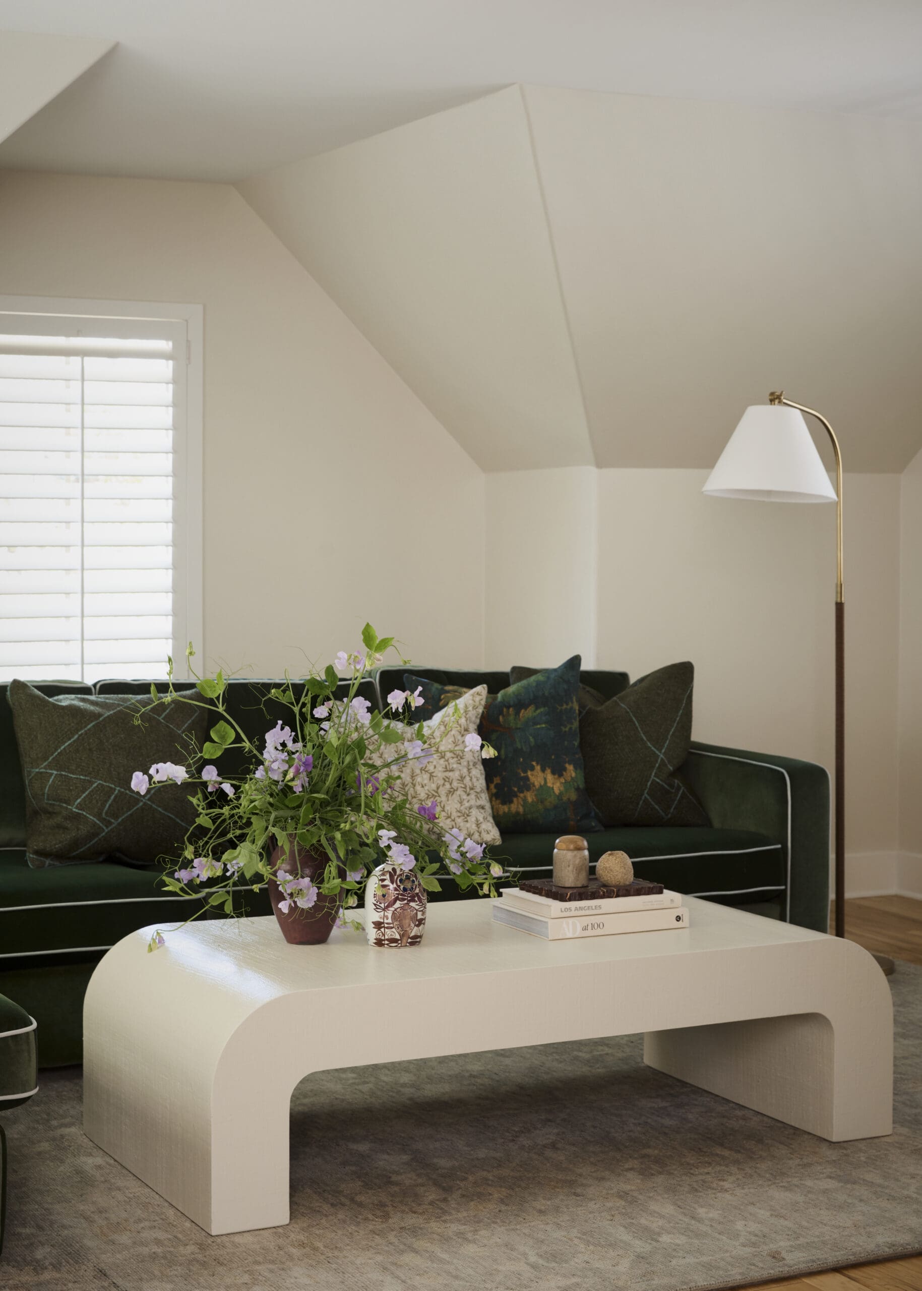 Living room scene with a curved white coffee table, dark green sofa, and a brass floor lamp; vase of purple flowers on the table and books nearby.