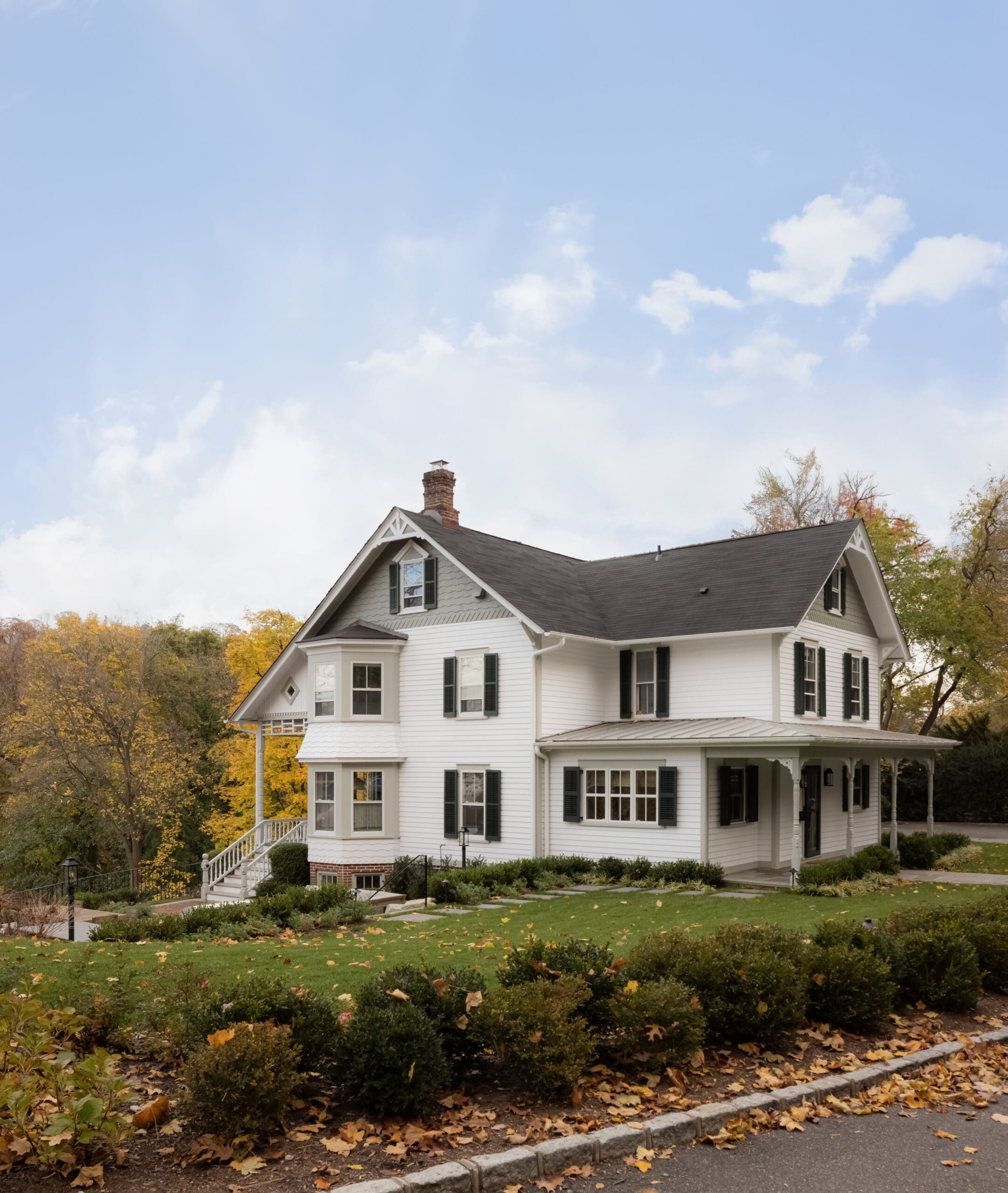 White two-story house with dark shutters and a wraparound porch, set among autumn trees and fallen leaves under a blue sky.