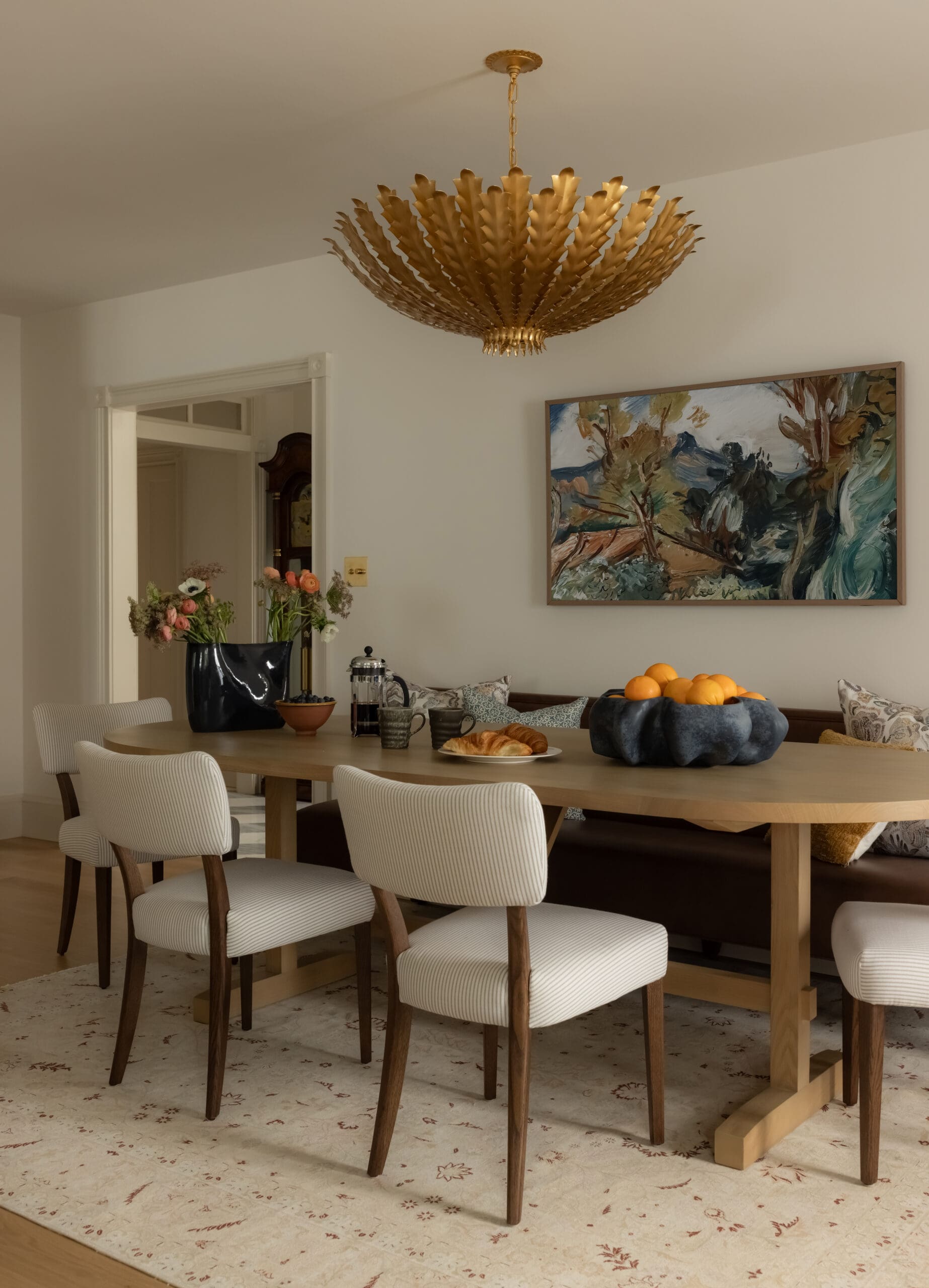 Elegant dining room with a large gold sculptural chandelier hanging above a wooden table and beige striped chairs.