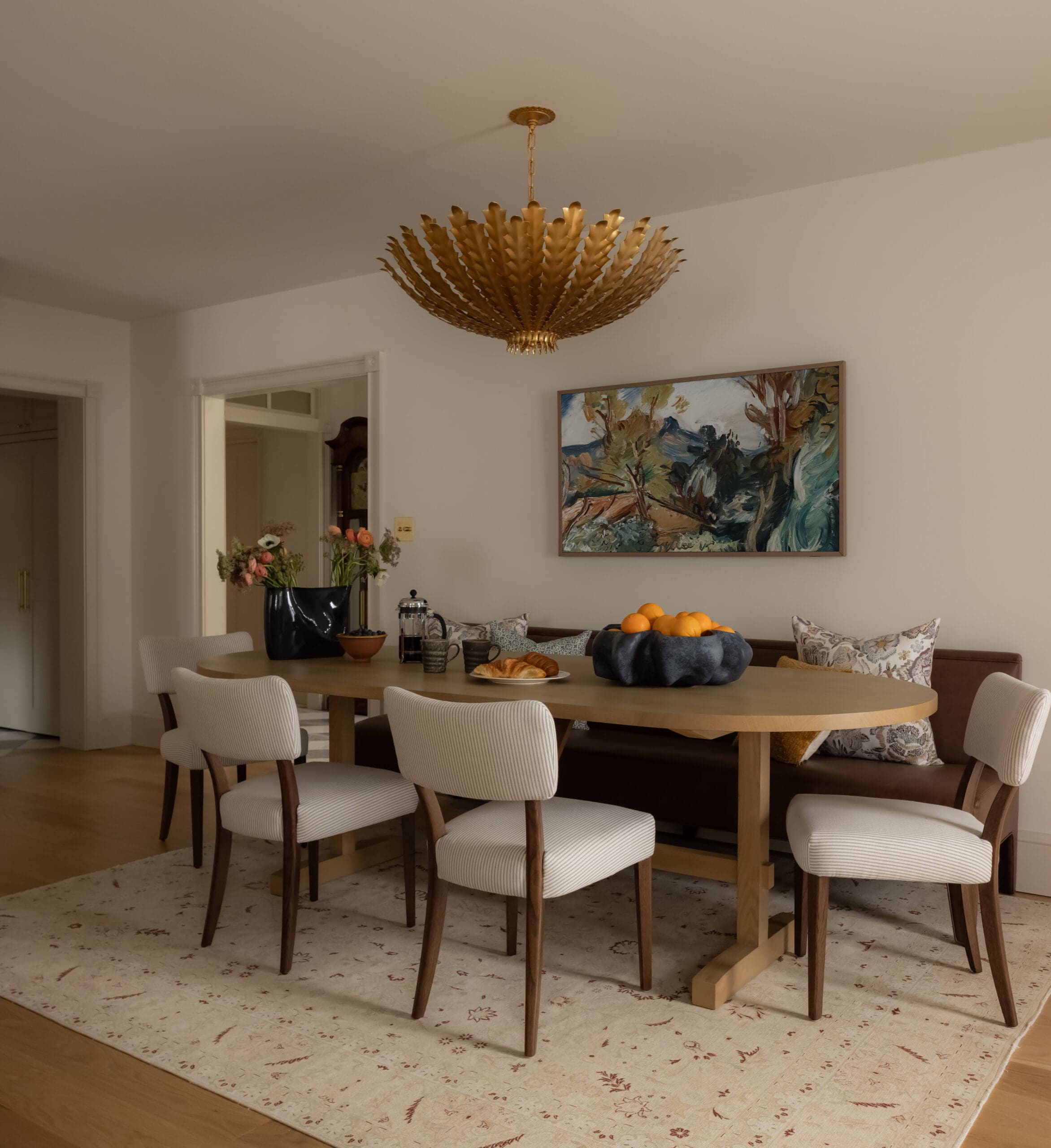 Gold sunburst chandelier hangs over a round wooden dining table with an orange centerpiece and cream chairs in a beige-walled dining room.