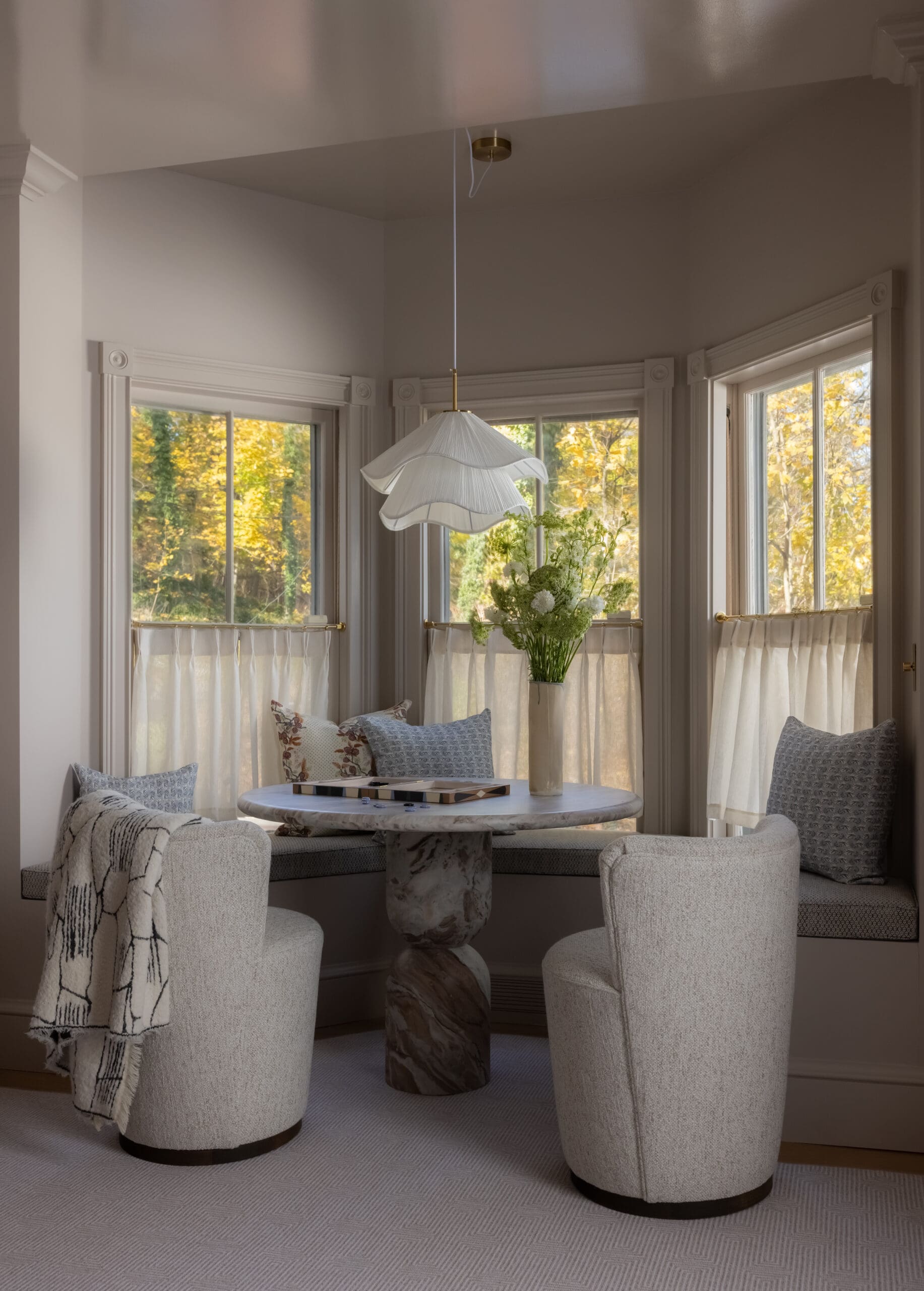 Cozy windowed dining nook with a round marble table, four cream upholstered chairs, a layered white pendant light, and a vase of white flowers; autumn trees outside.