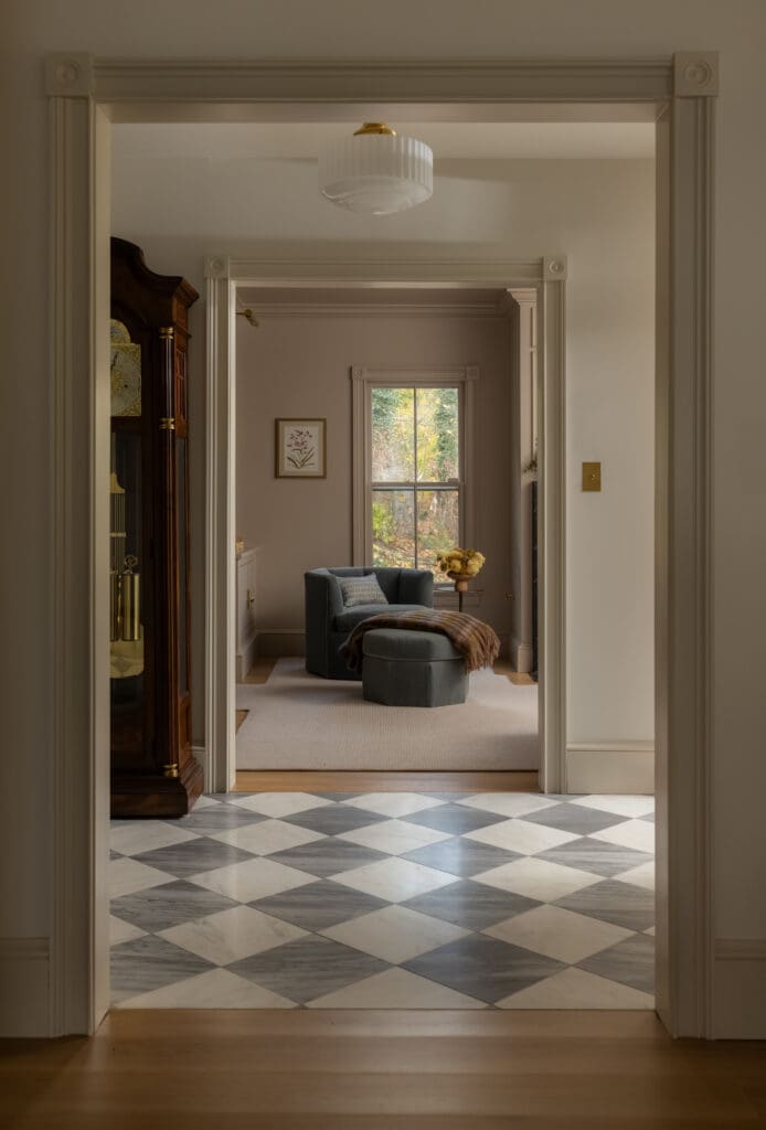 A view through a sequence of doorways into a warmly lit living room with a blue chair, ottoman, and window showing fall foliage outside.