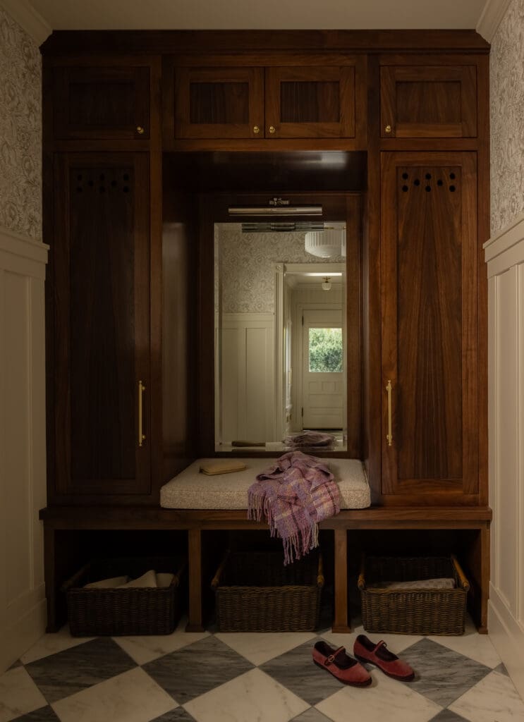 Dark wood built-in wardrobe with a full-length mirror, bench seating, and three woven baskets beneath; purple scarf and red shoes on a checkered marble floor.