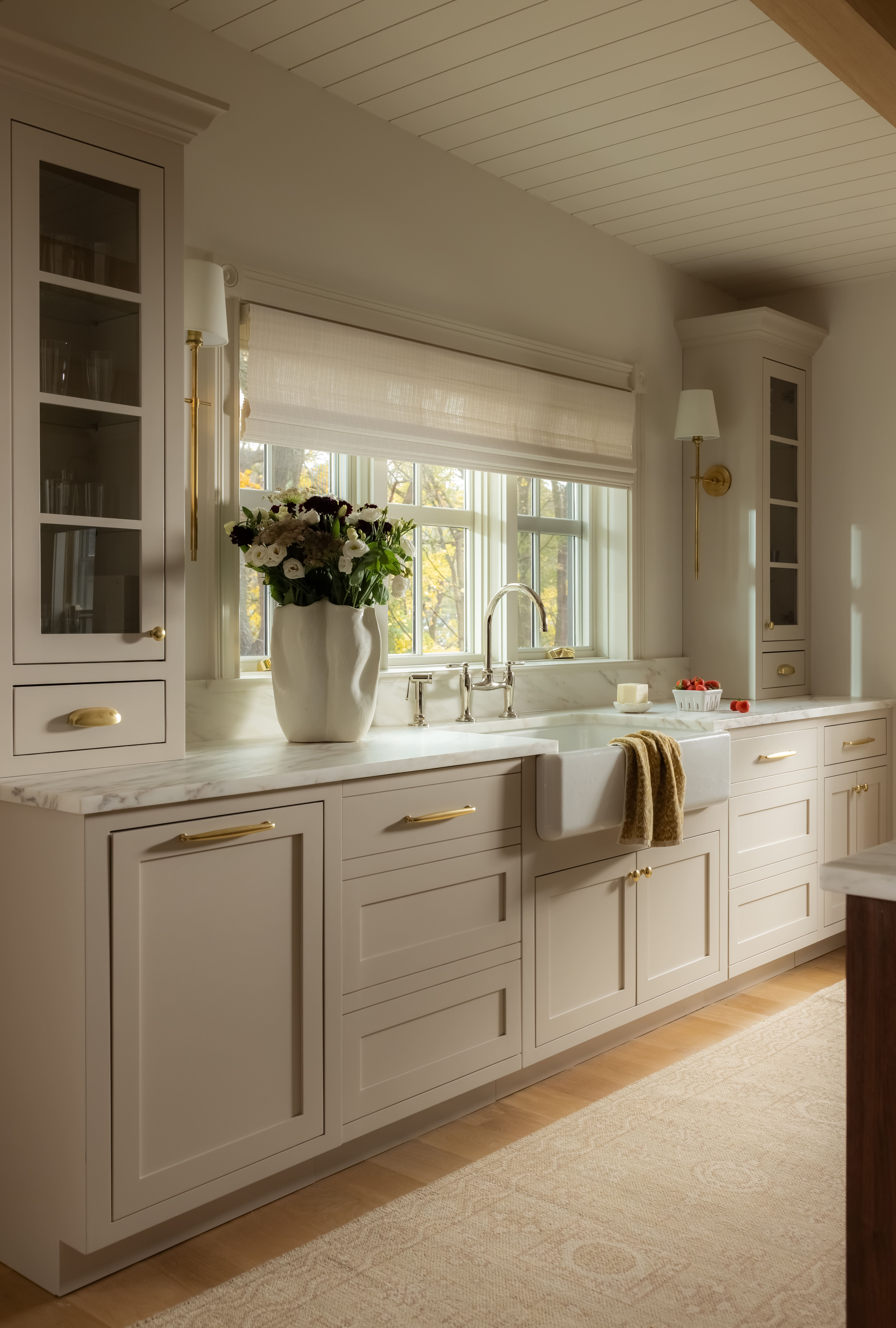 Cream kitchen with marble countertops, white sink under a window, and gold cabinet hardware.