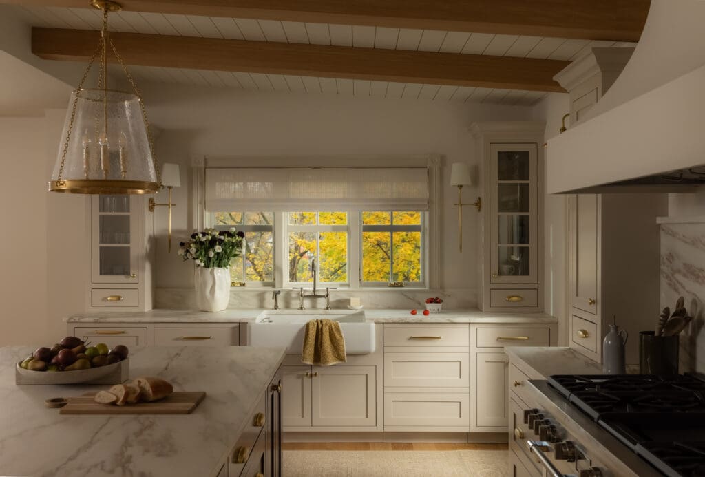 Bright white kitchen with marble countertops, brass hardware, and a large glass pendant light hanging from wooden beams.
