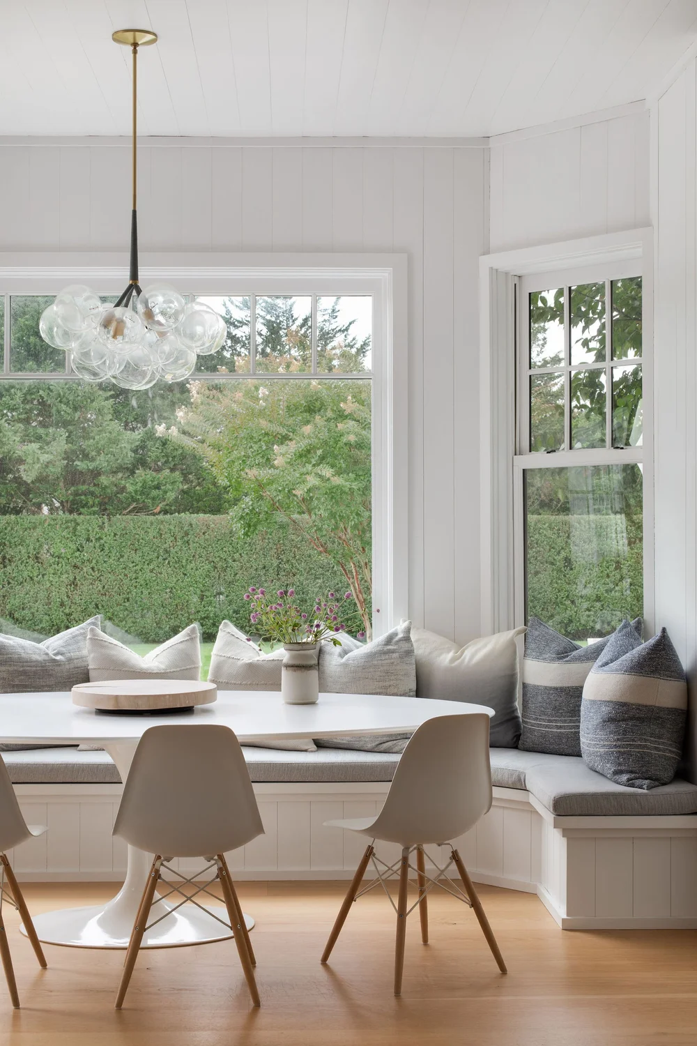 Bright dining nook with a white round table, modern chairs, a glass globe chandelier, and a window seat with cushions overlooking a garden view.