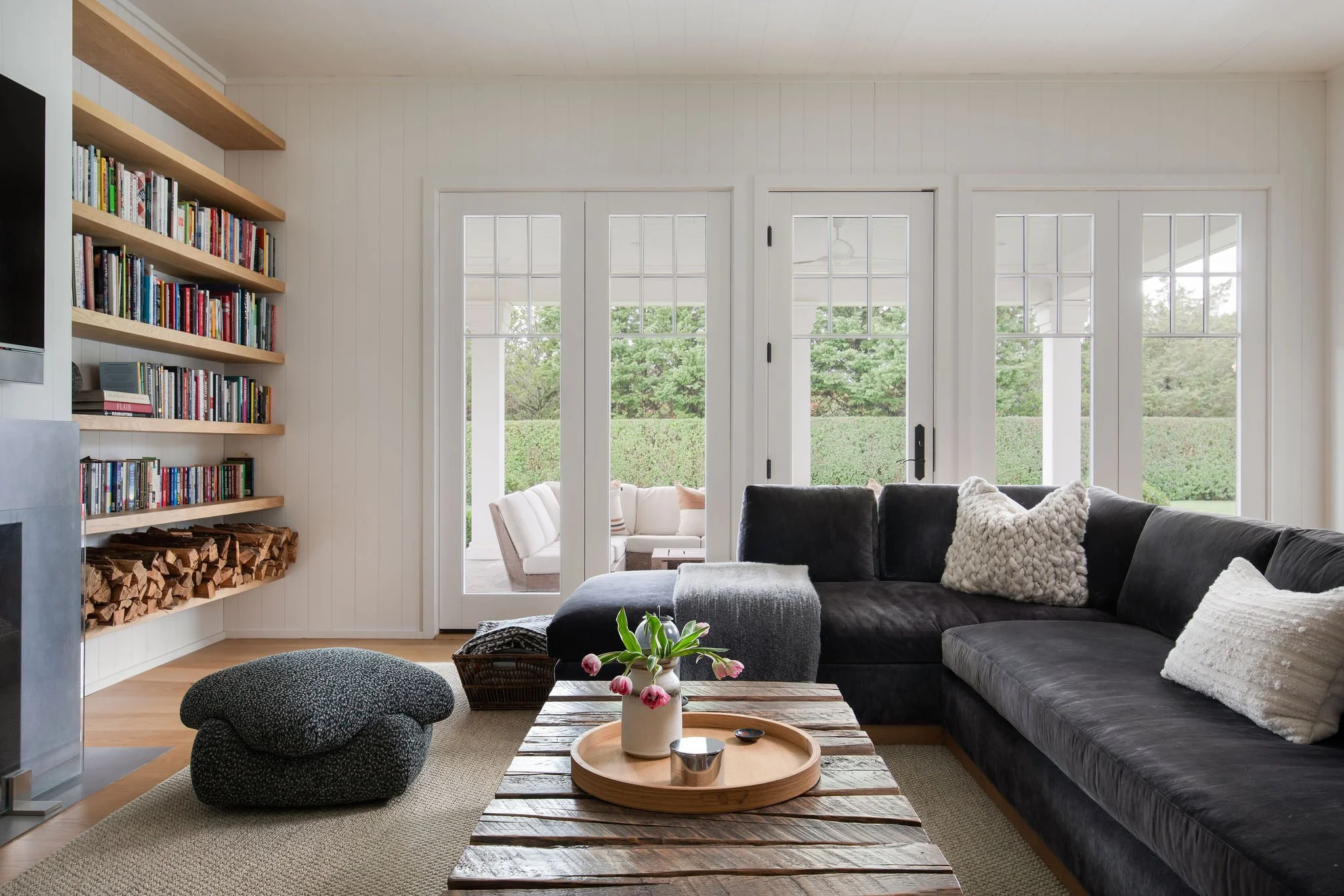 Bright living room with glass French doors, a dark sectional sofa, fluffy pillows, and a wooden coffee table; bookshelves to the left.