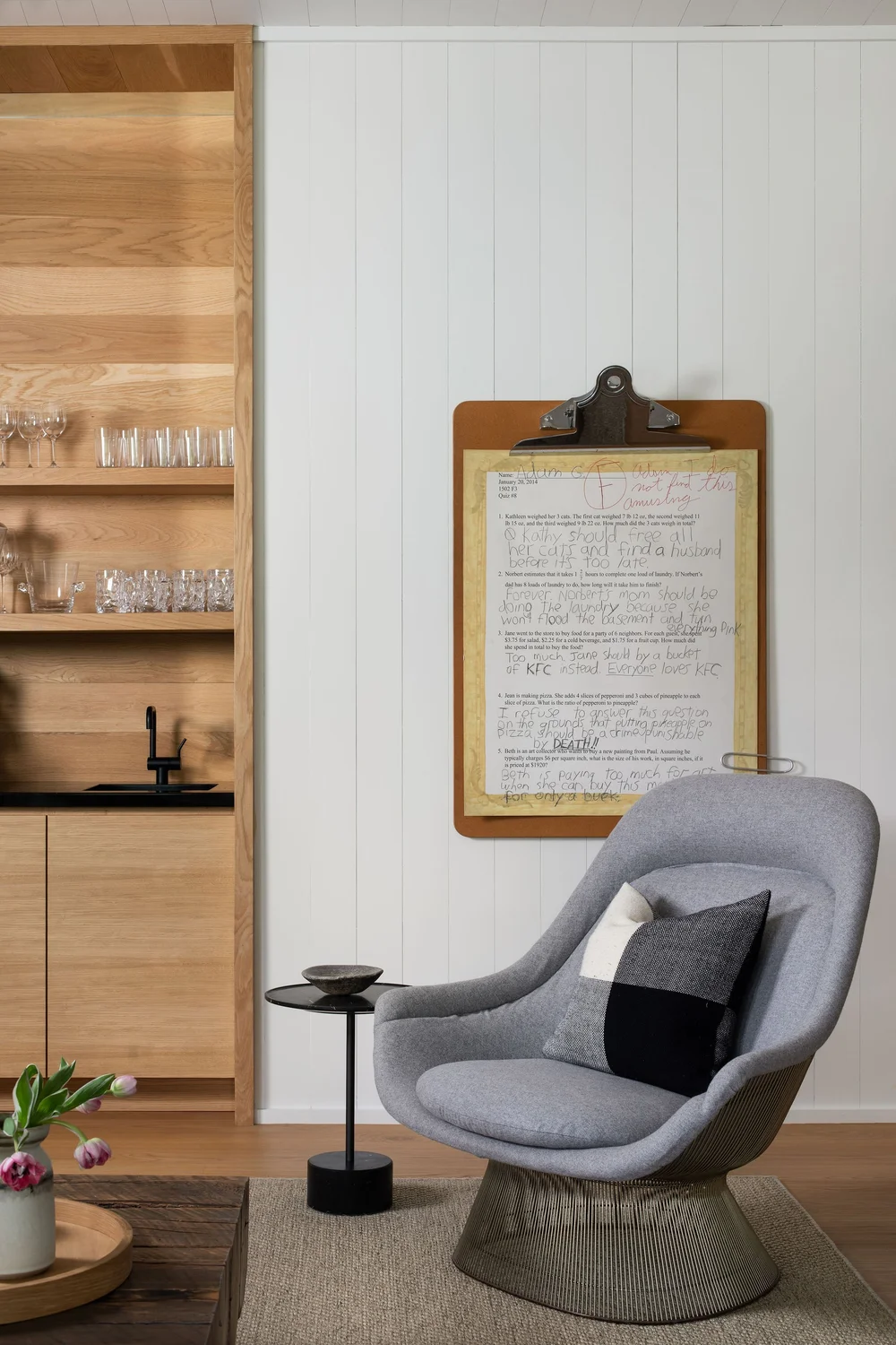 Grey curved lounge chair with a metal pedestal base, a small black side table, and a vase of pink tulips on a wicker tray in a cozy living corner; white paneled wall features a clipboard with papers.