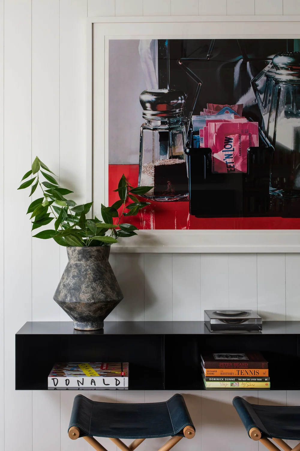 Potted green plant in a weathered ceramic vase sits on a black console under a framed still life photograph.